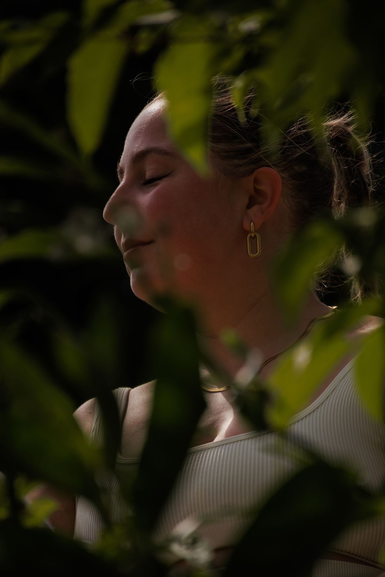 Close-up portrait through leaves showing profile view with soft natural lighting and bokeh effect.