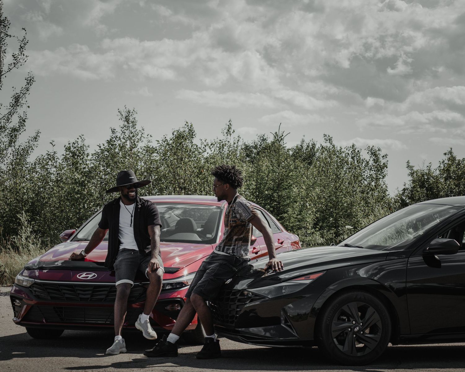 Two people leaning against black cars parked on a rural road with trees in the background.