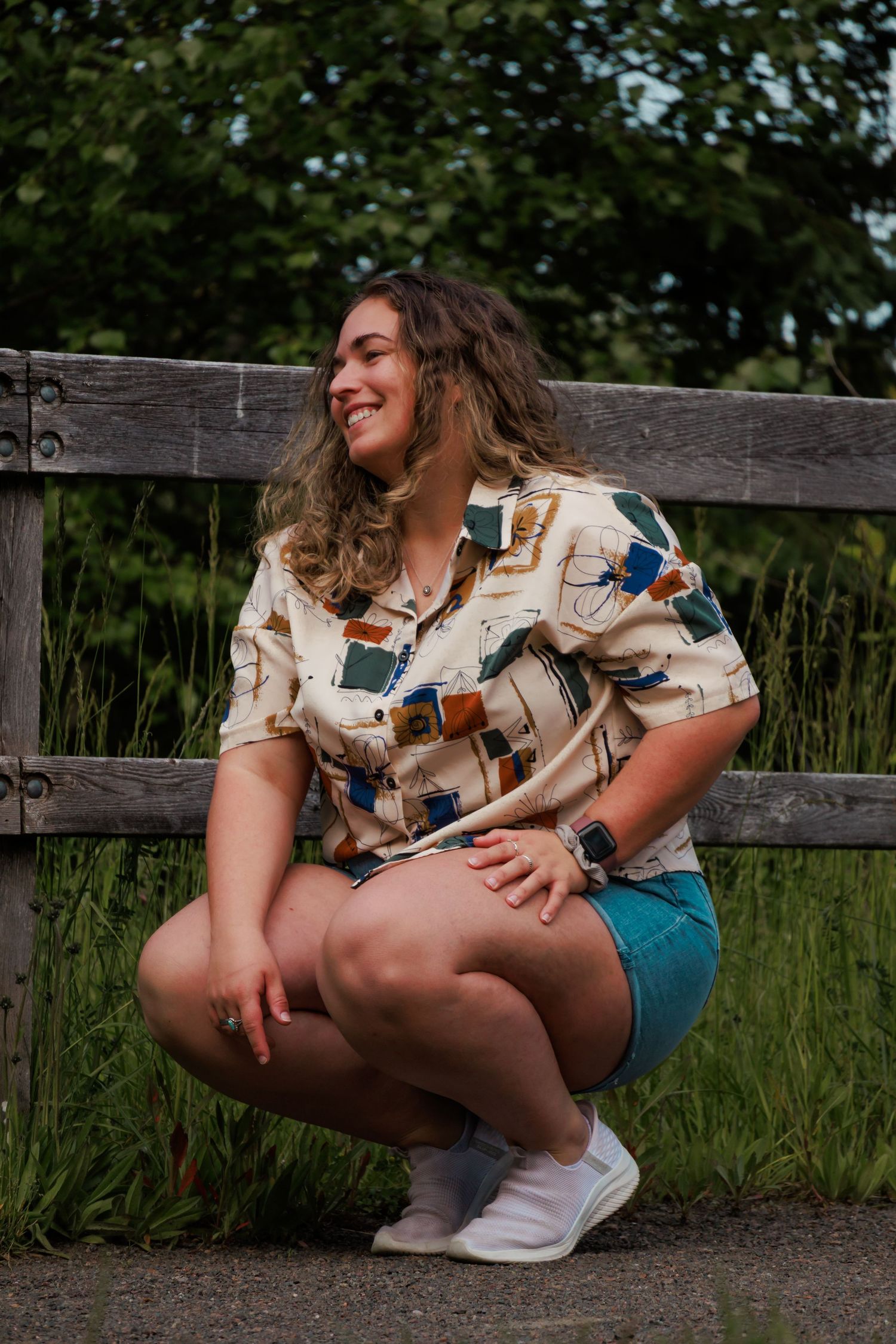 Person in patterned shirt and denim shorts sitting casually on wooden bench in outdoor nature setting.