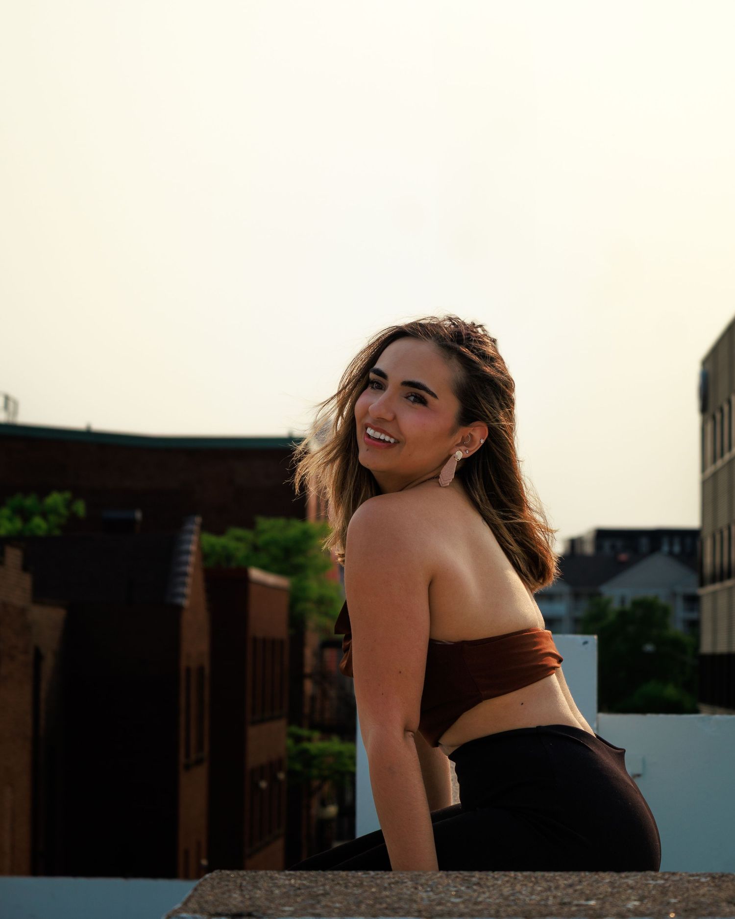 Person in black dress posing at sunset against stone wall with silhouetted buildings in background.
