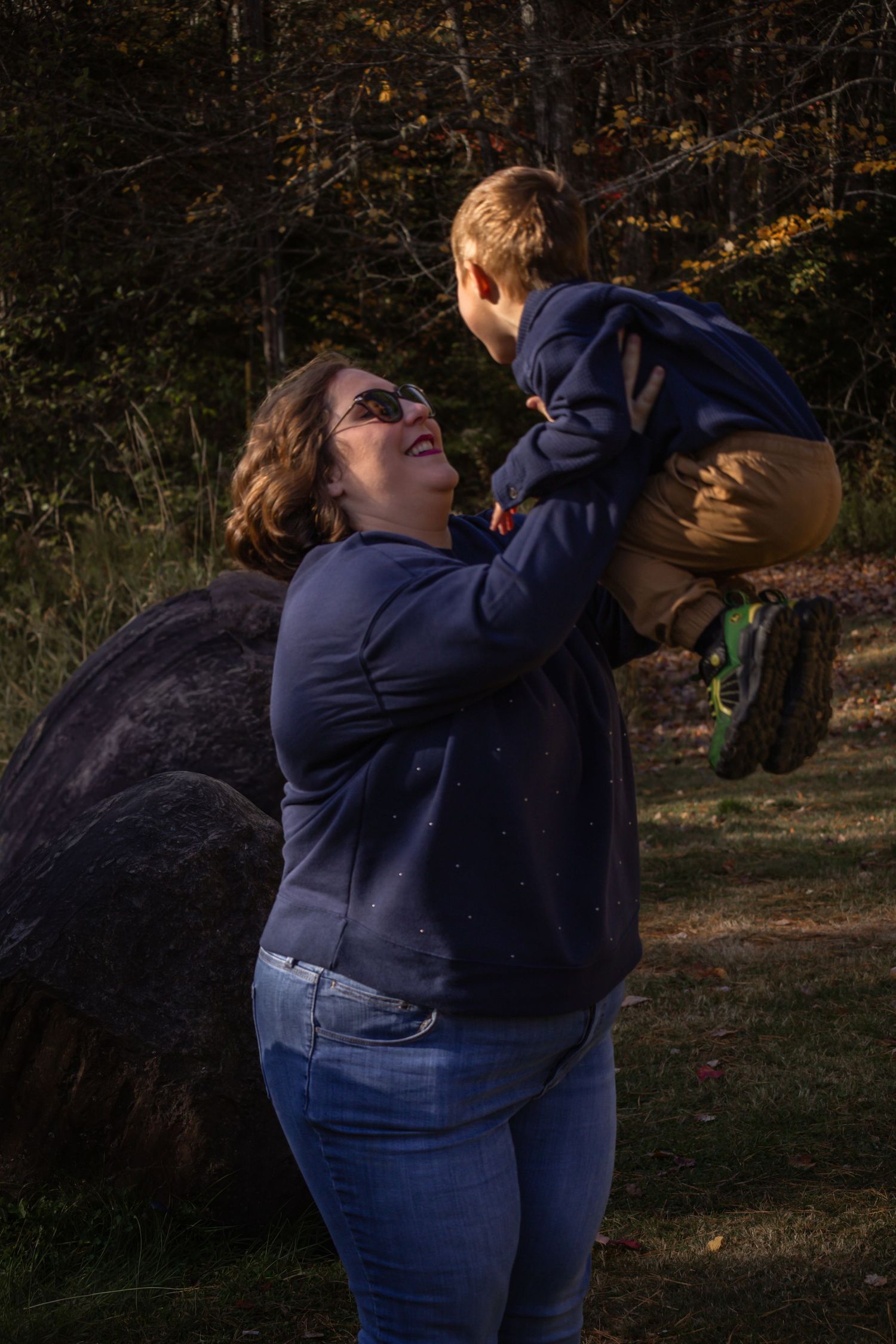 A joyful moment captured outdoors as a parent lifts a small child in the air against an autumn forest backdrop.