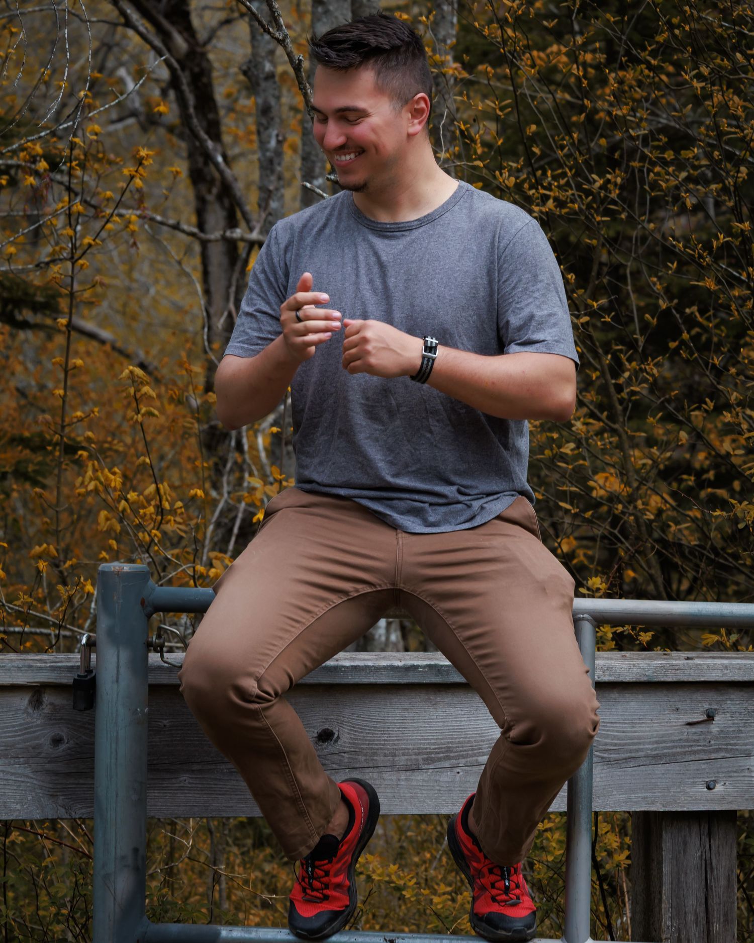 Person in grey shirt and khaki pants sitting casually on wooden railing with fall foliage behind.