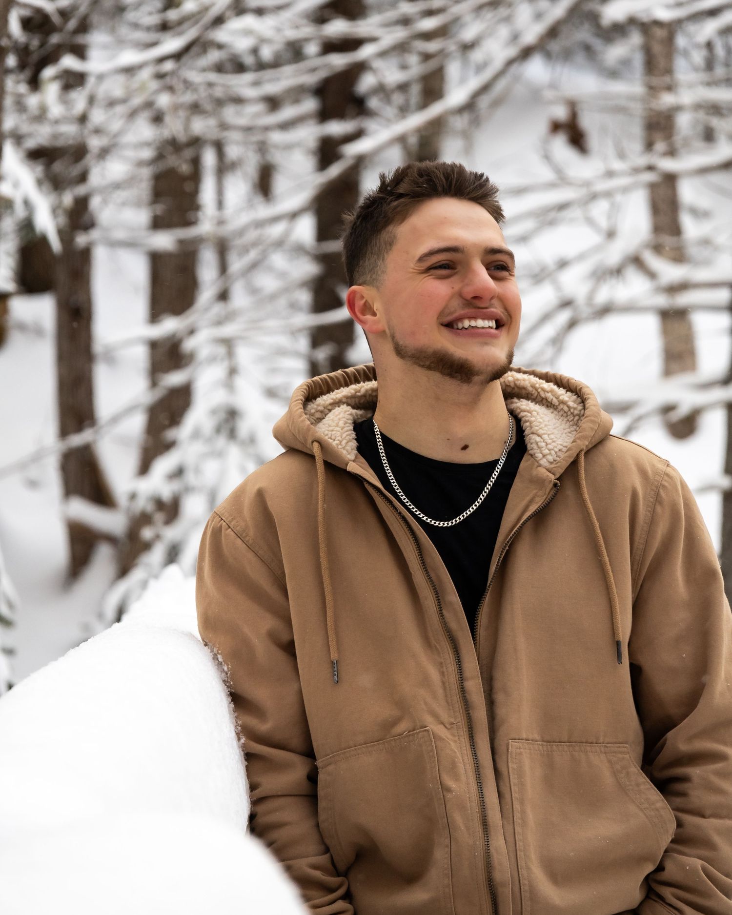 Person in tan hoodie and black turtleneck smiles against snowy winter forest background.