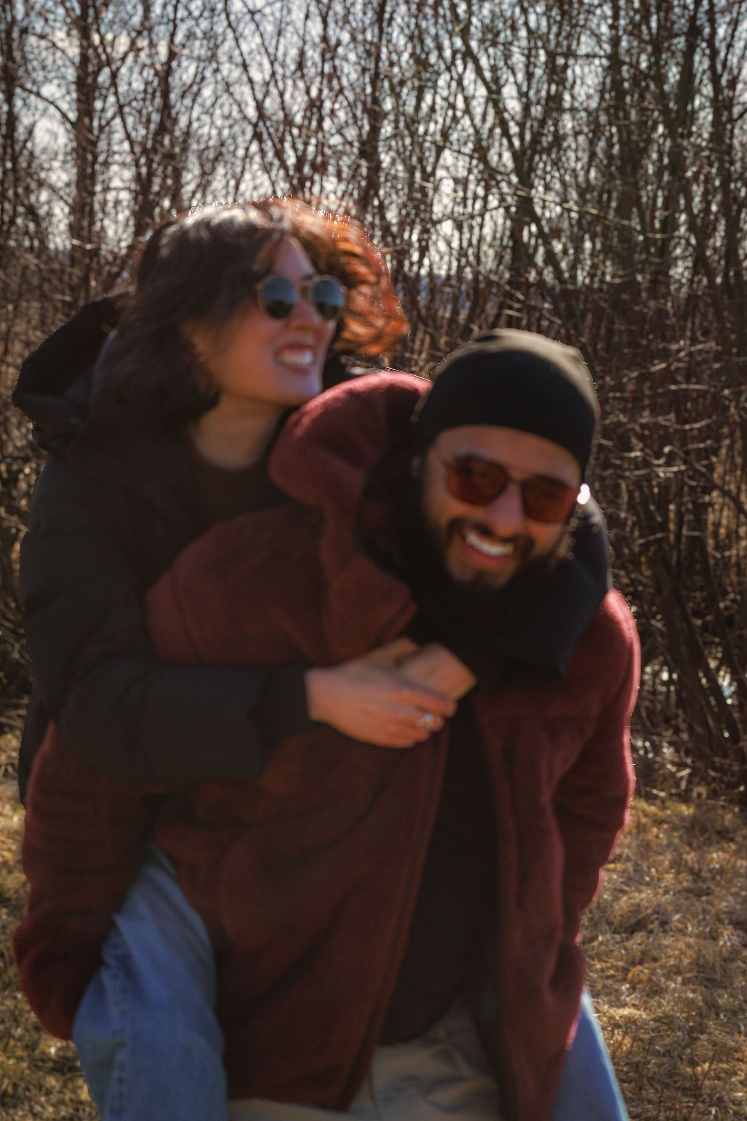 Couple in sunglasses sharing a playful moment outdoors among bare autumn trees.