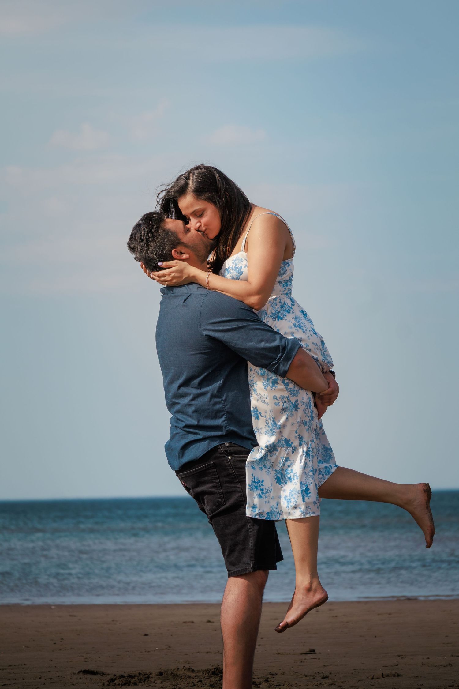 Romantic couple in playful embrace on beach with blue sky and ocean backdrop.