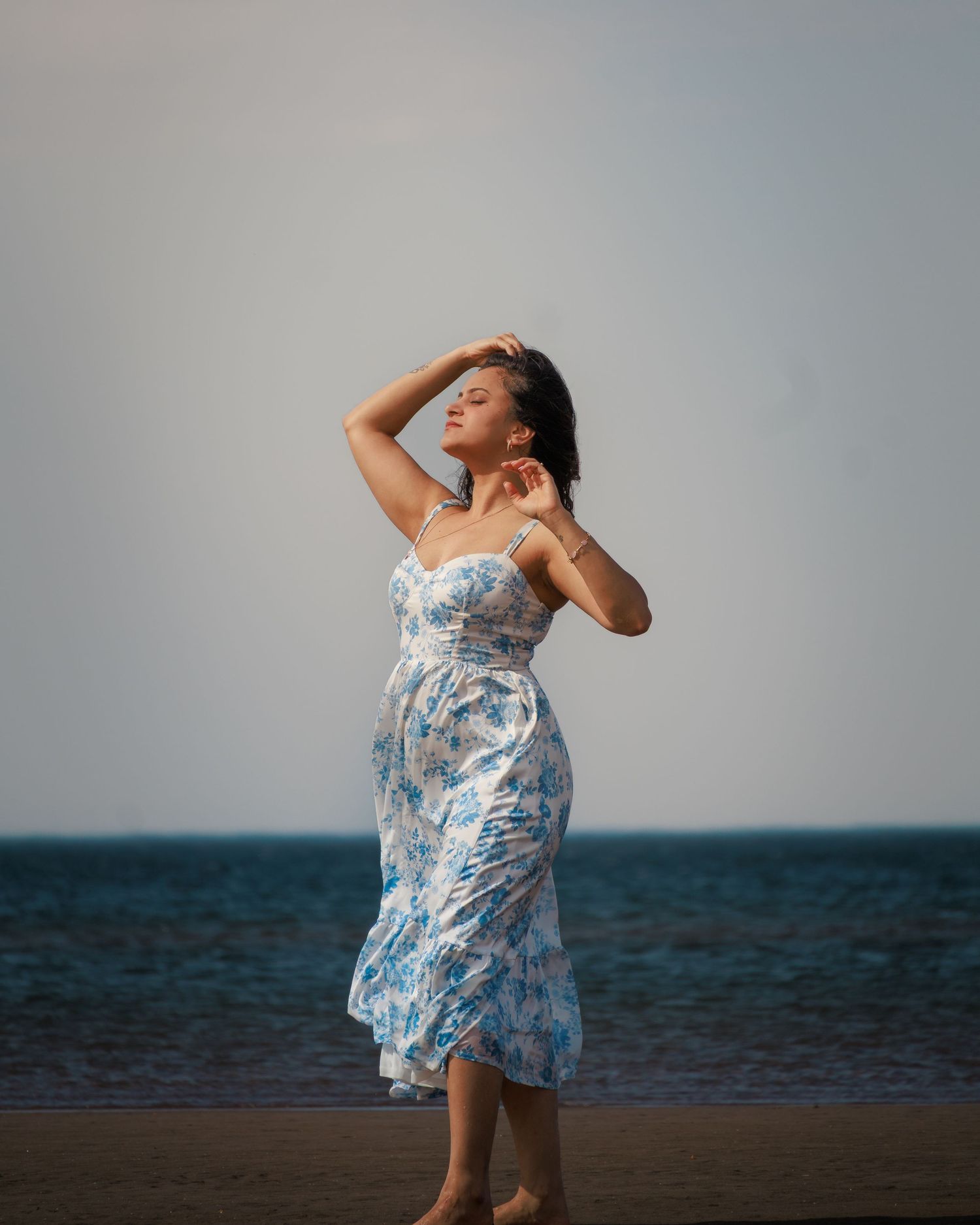 Person in flowing blue floral dress dancing expressively on beach against ocean horizon.