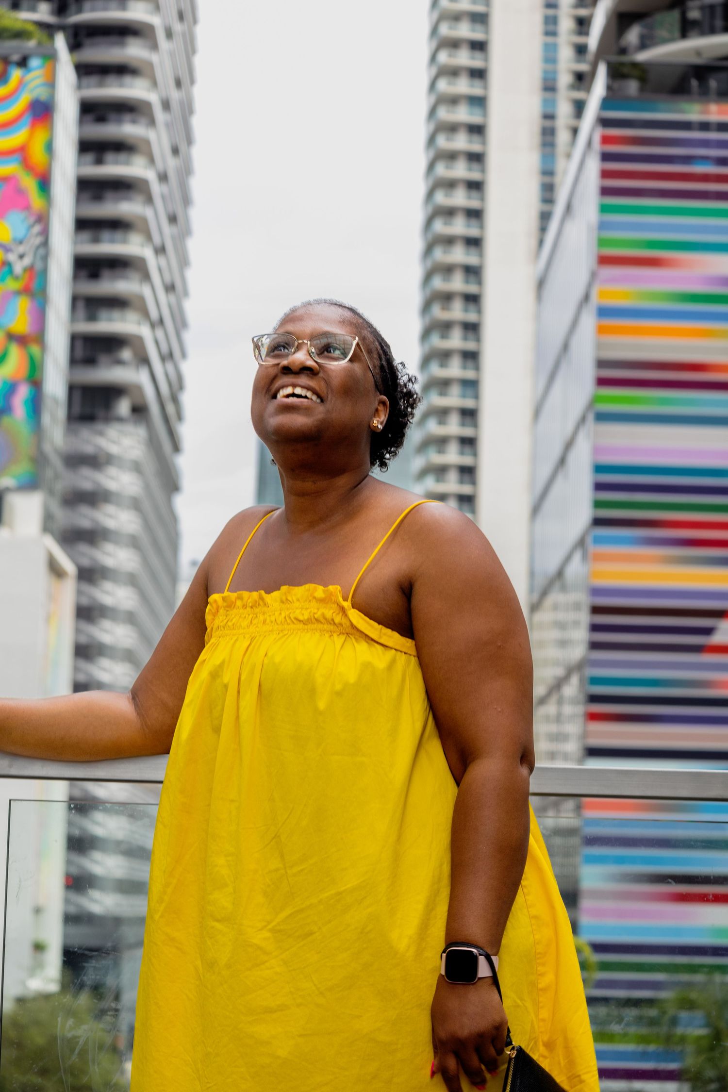 Woman in bright yellow dress laughs joyfully against urban backdrop with colorful striped building.