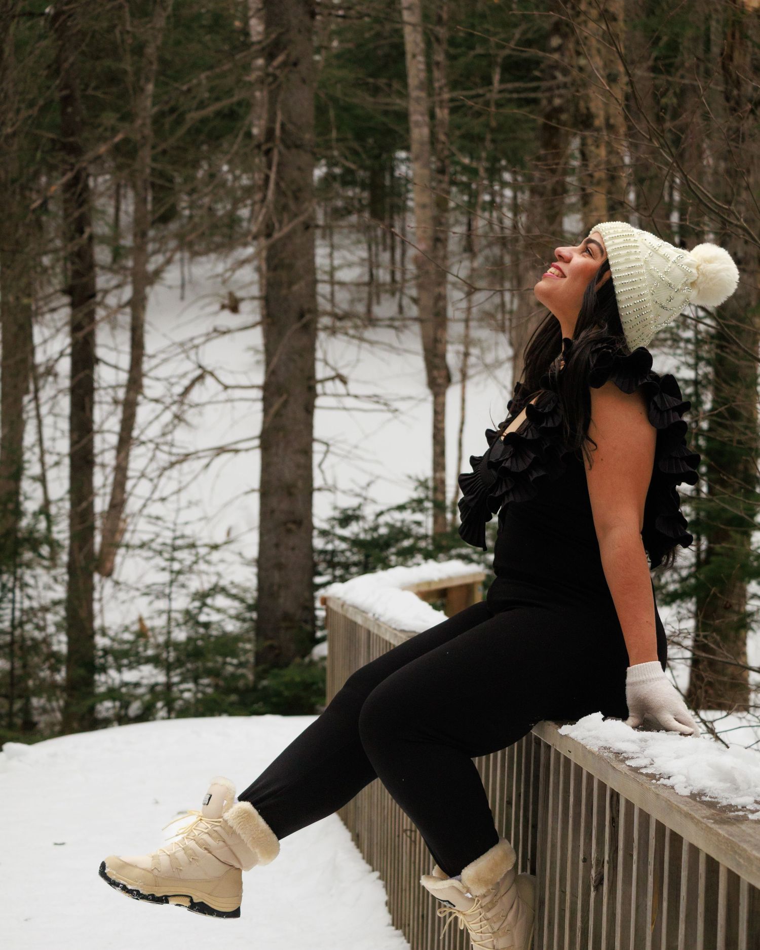 Person in black outfit sitting on snowy railing in winter forest setting.