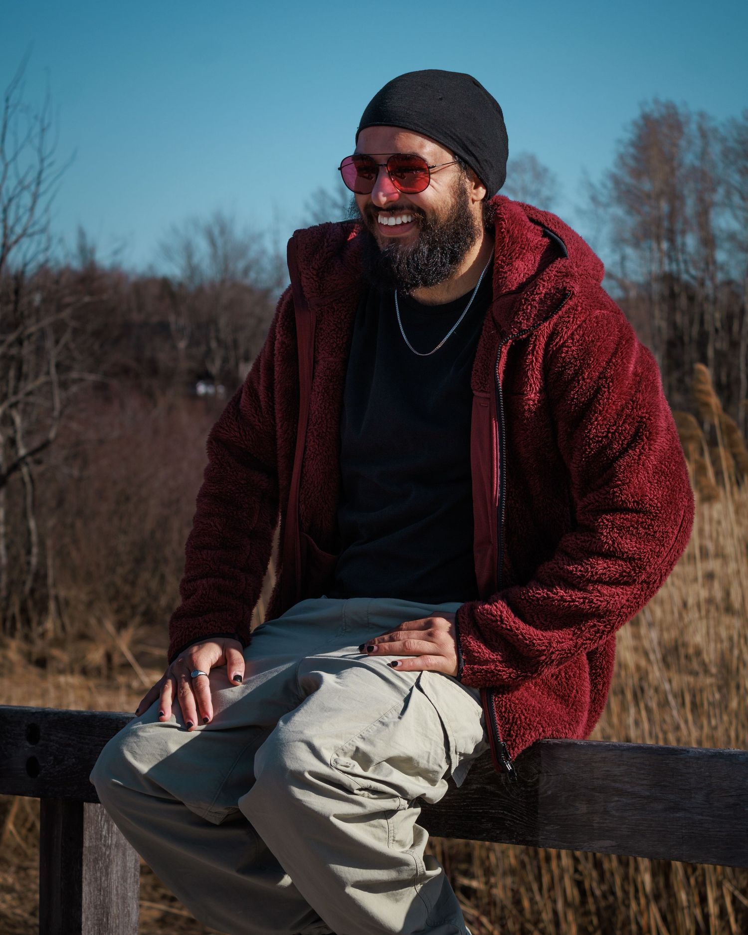 Person in red fleece jacket and beanie sitting casually on wooden railing against winter forest backdrop.