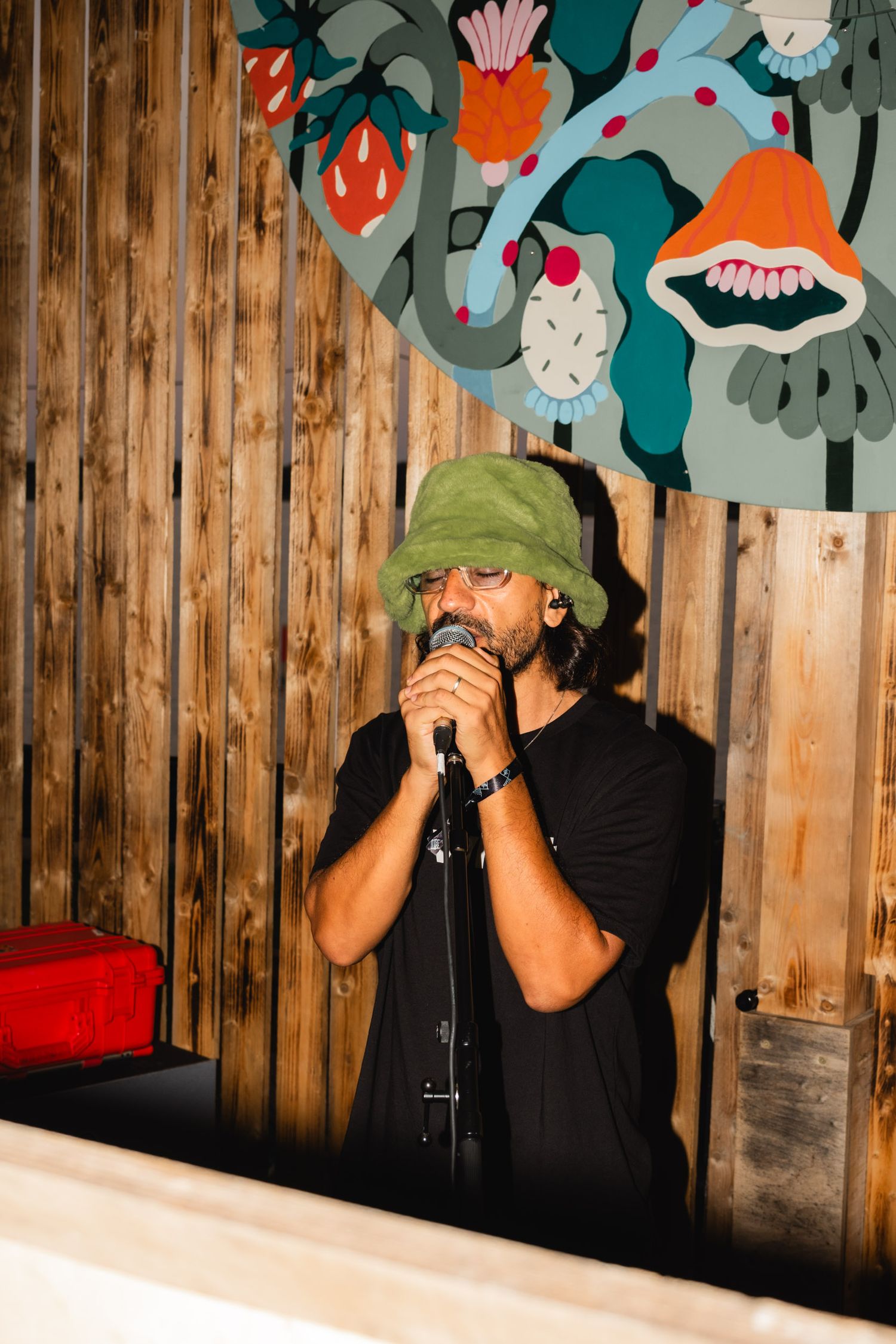 Performer in a green hat singing into microphone against wooden wall with colorful art.