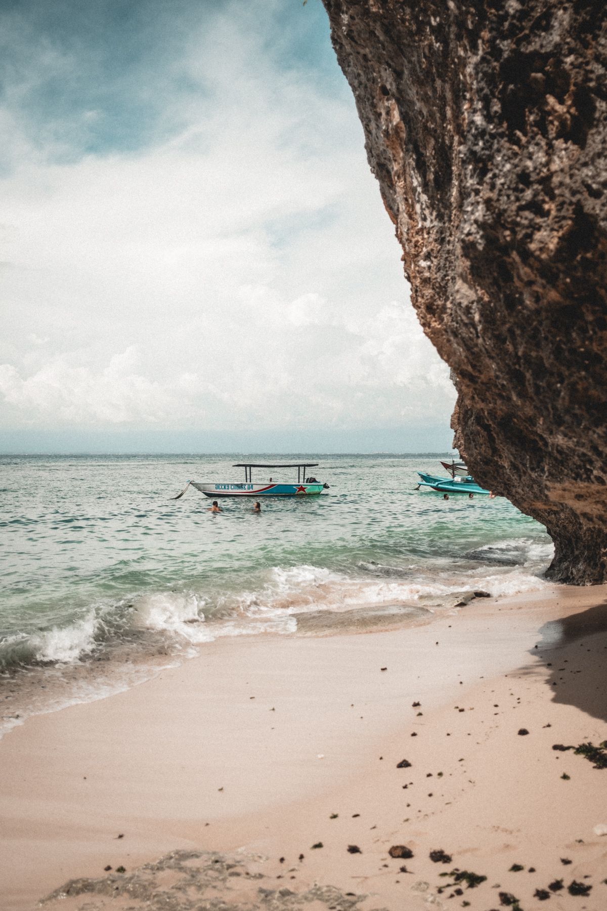 Small boats anchored near dramatic limestone cliff cave overlooking turquoise ocean waters.