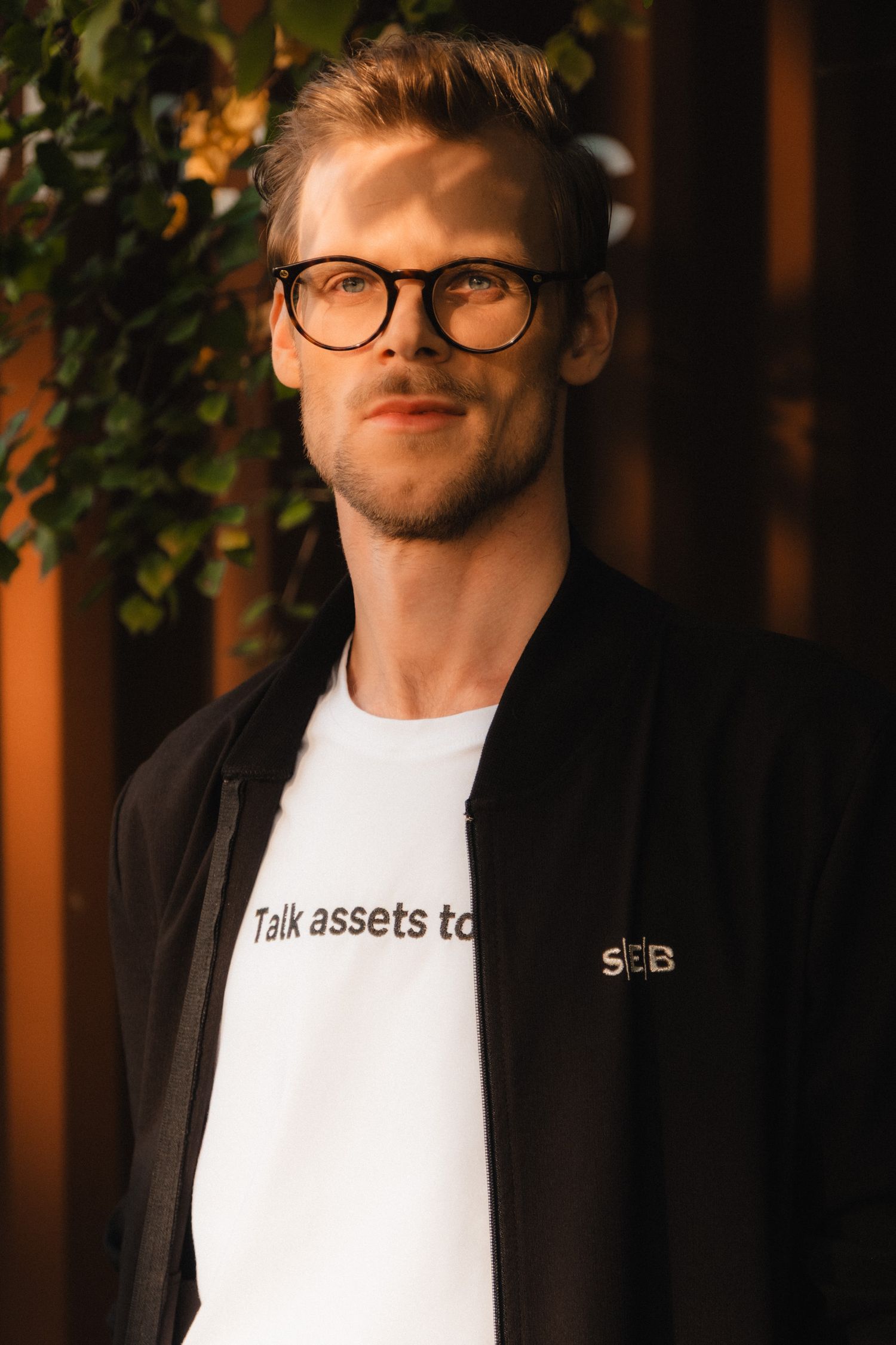 Person wearing glasses and a white t-shirt poses against dark background with green foliage visible.