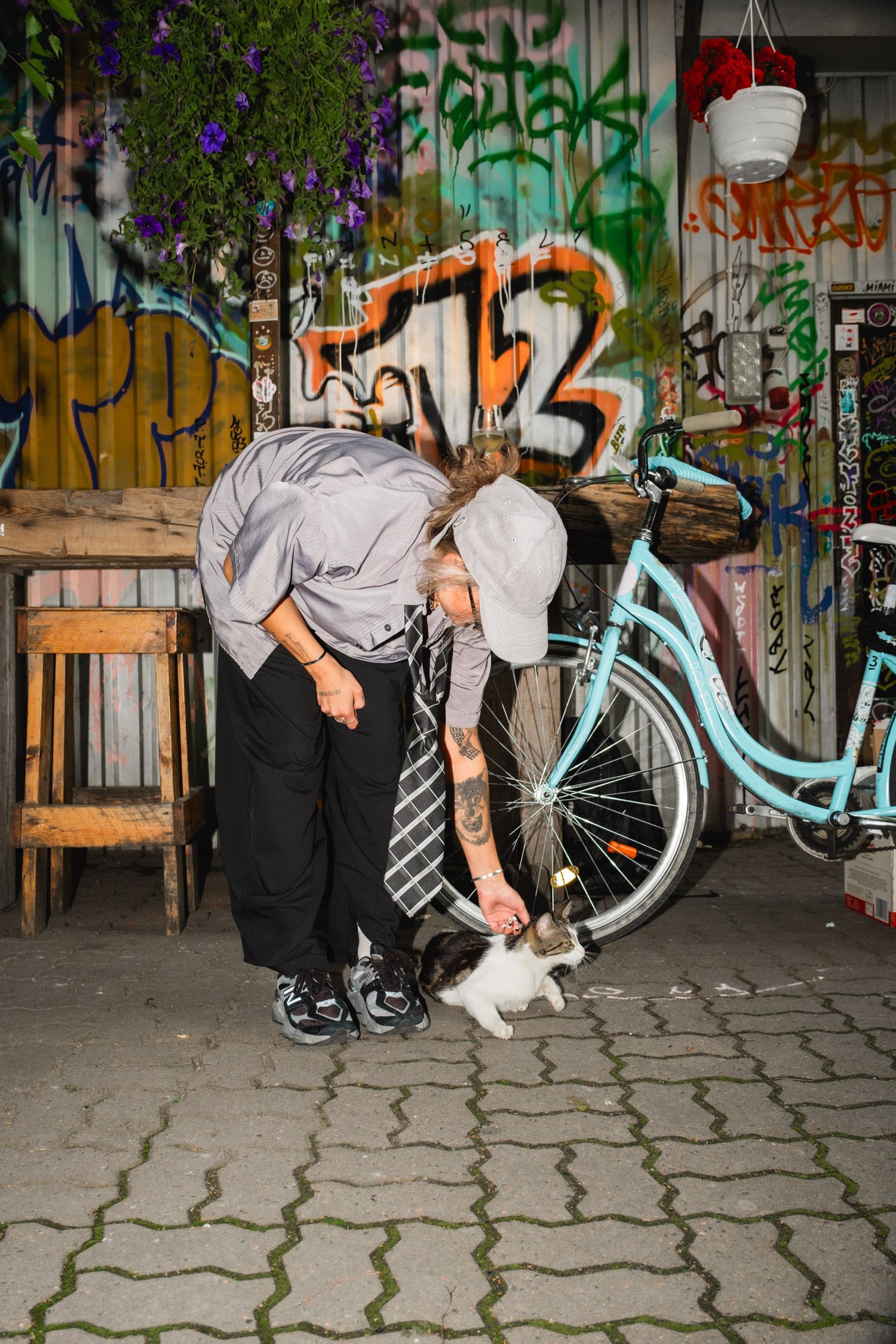 Someone in casual attire pets a cat near a light blue bicycle and graffiti wall.