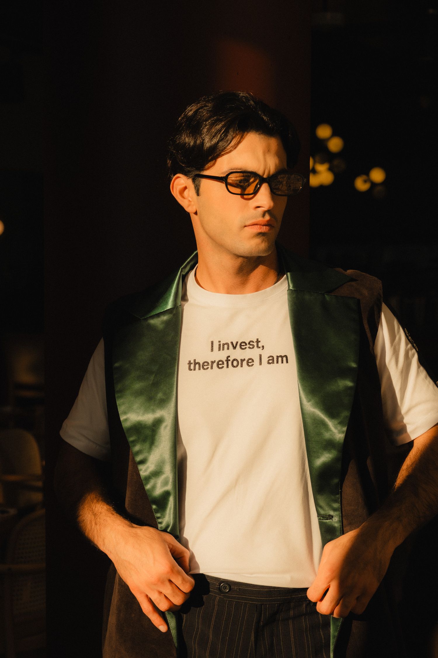 Person wearing glasses and a green vest over a white t-shirt with text 'I invest, therefore I am' in dark lighting.