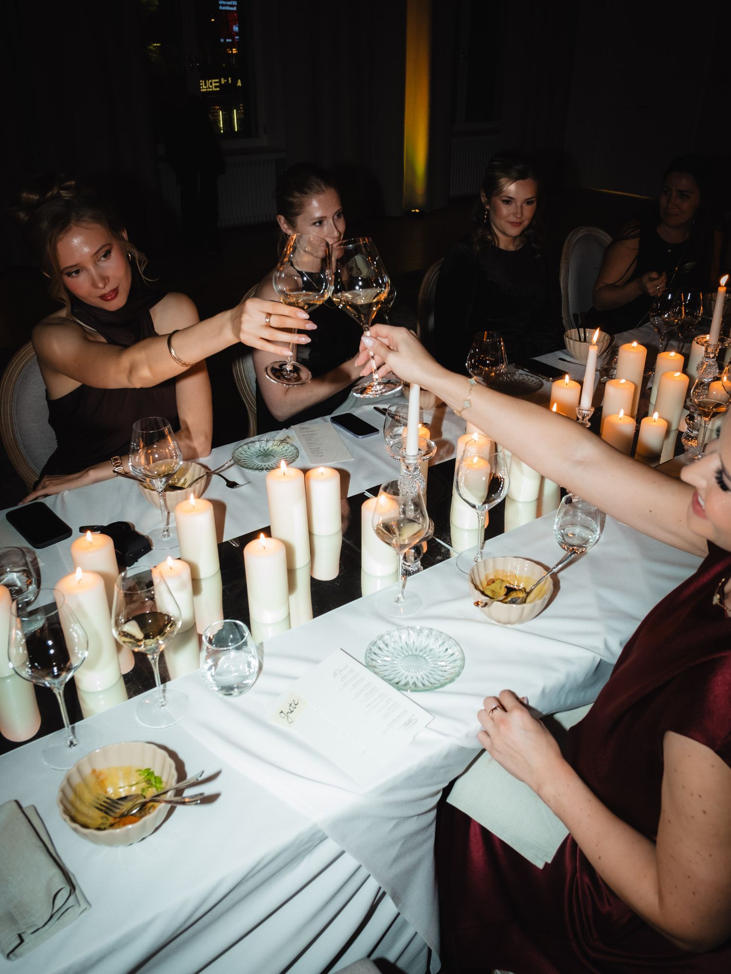 Dinner party guests toasting with wine glasses around a candlelit table with white tablecloth.