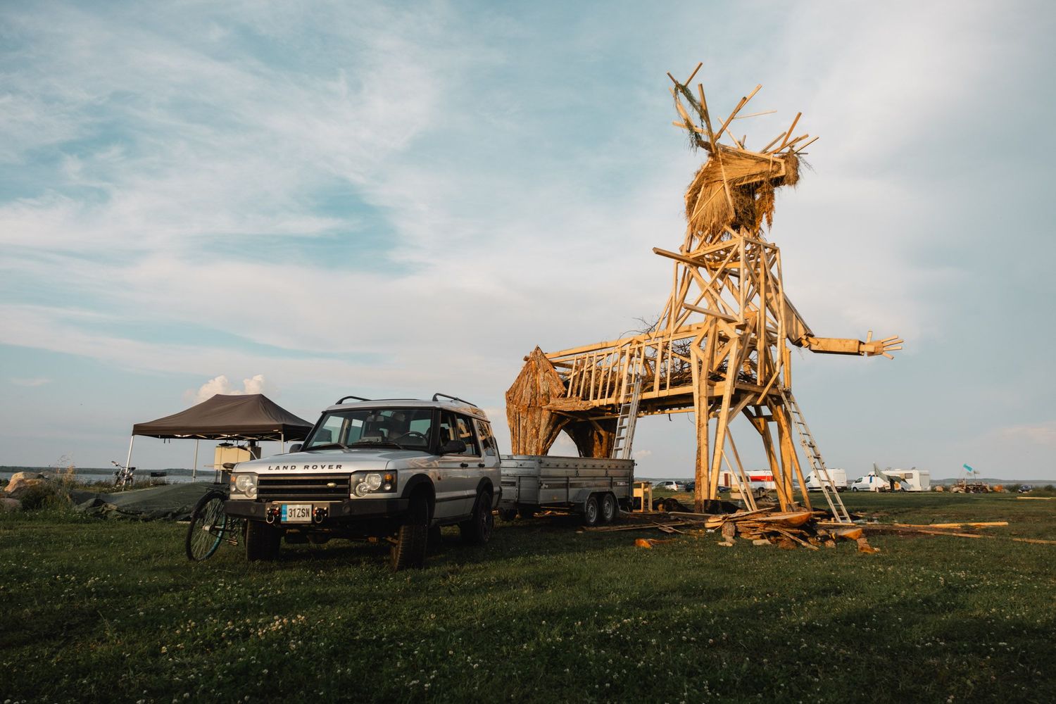 Large wooden deer sculpture installation next to parked car in rural field.