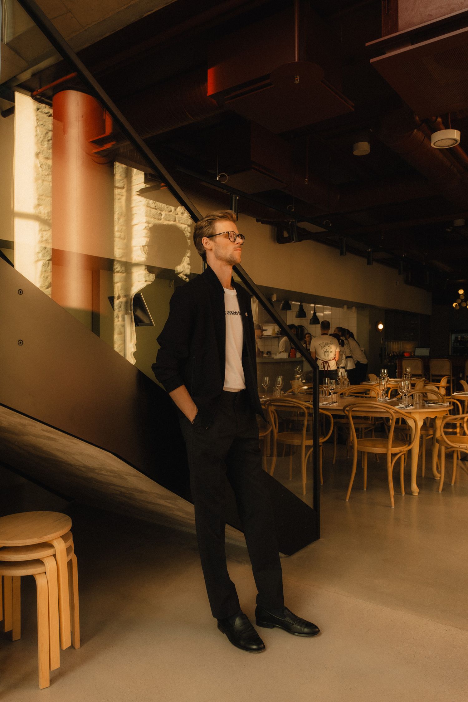 A person in business attire stands in a modern restaurant interior with wooden furniture and warm lighting.