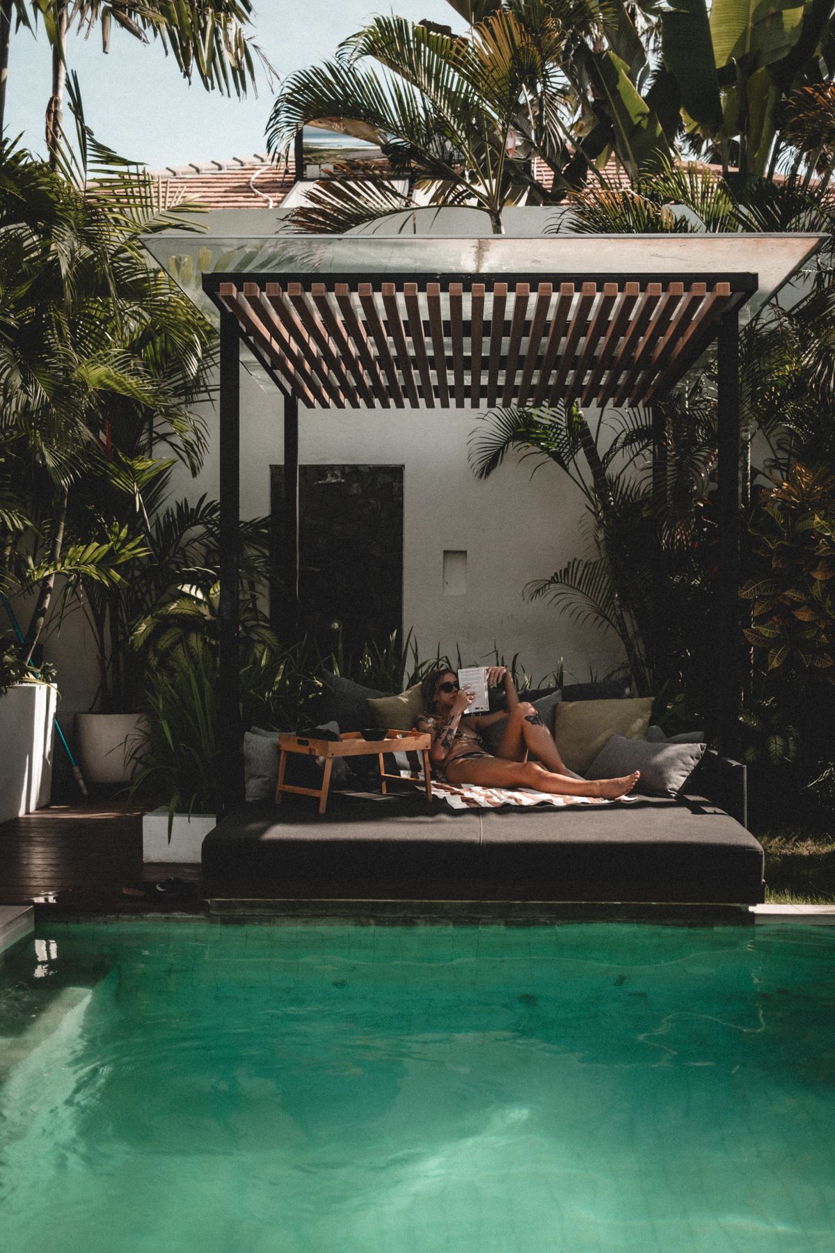 Luxurious poolside cabana with wooden pergola and comfortable daybed surrounded by tropical plants.