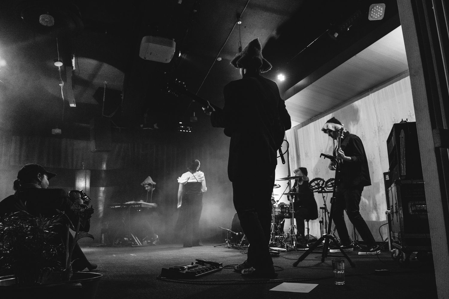 Wide angle concert venue shot showing performers and stage setup in moody black and white lighting.