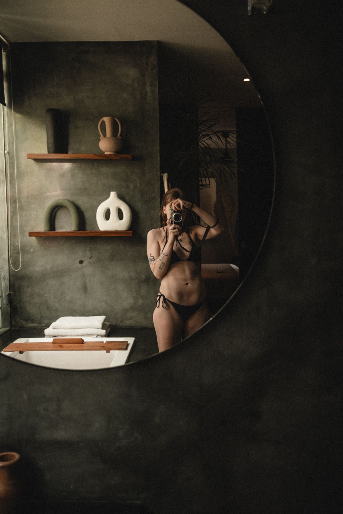Dark moody bathroom interior with round mirror and decorative shelves displaying ceramic vessels.
