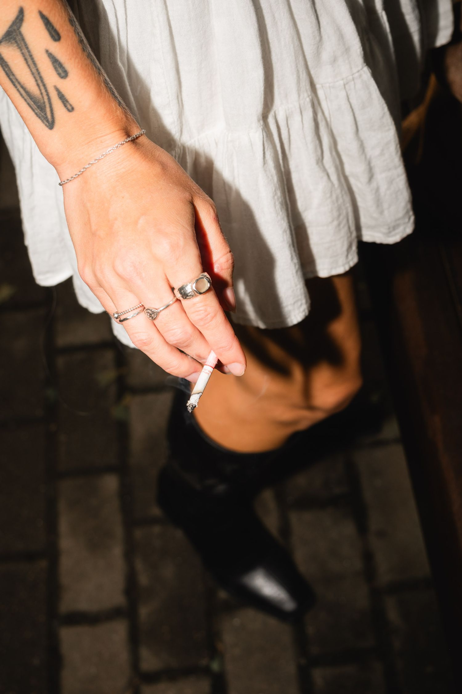 A tattooed hand with rings holds a cigarette against a white dress background.