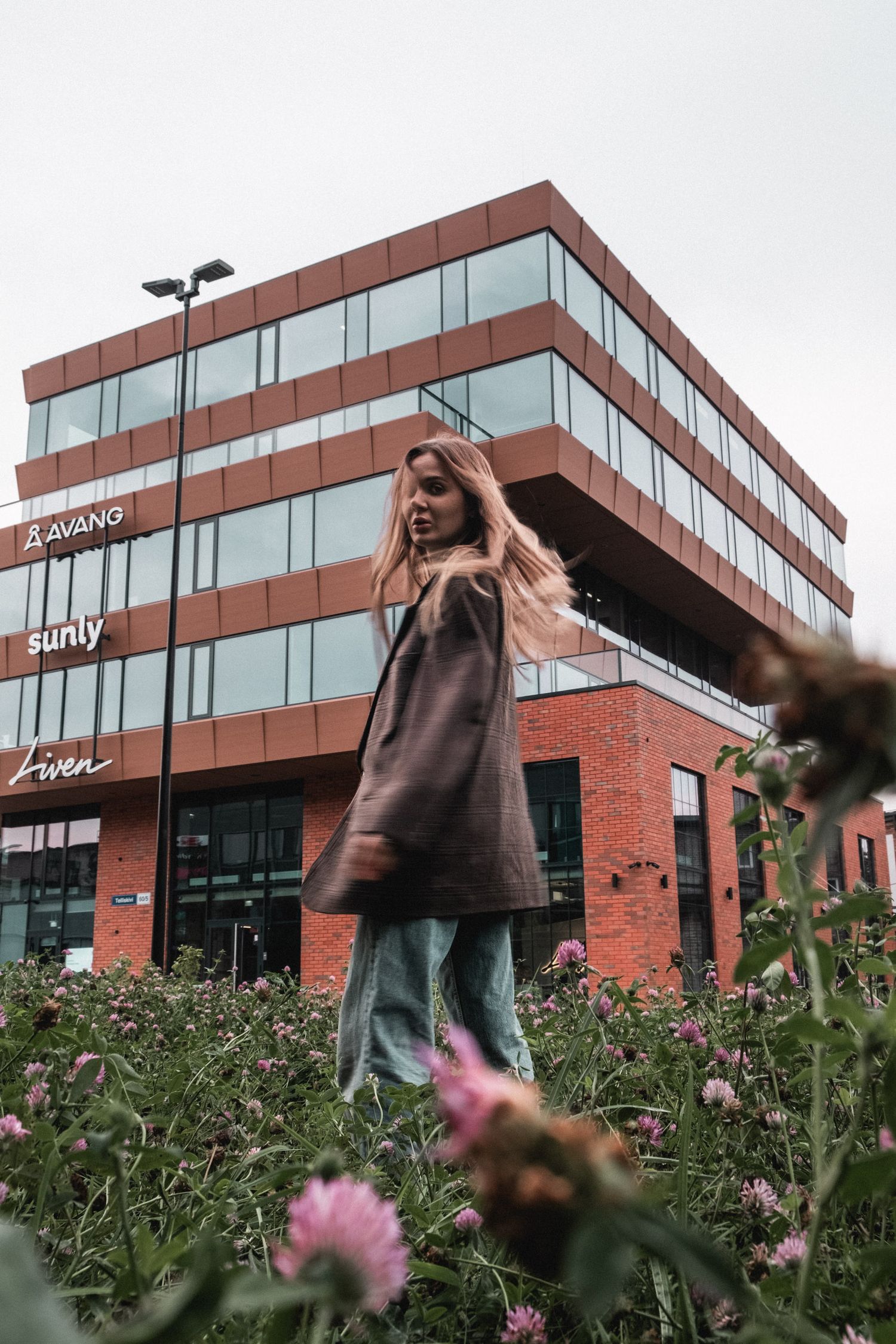 Figure in brown coat dancing in front of red brick building with pink flowers in foreground.
