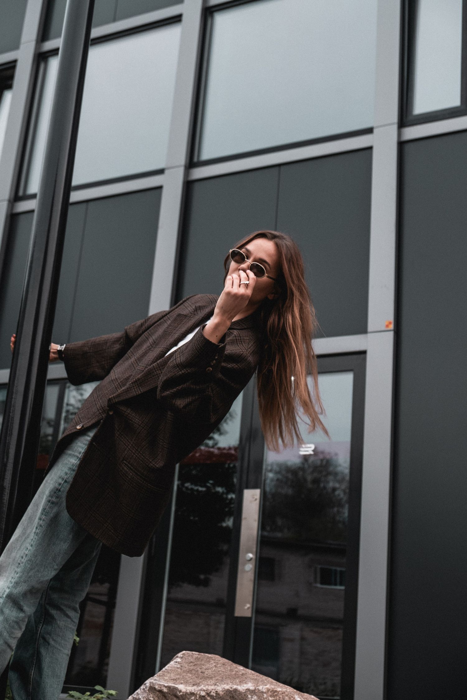 Person in brown jacket leaning back against modern glass building exterior.