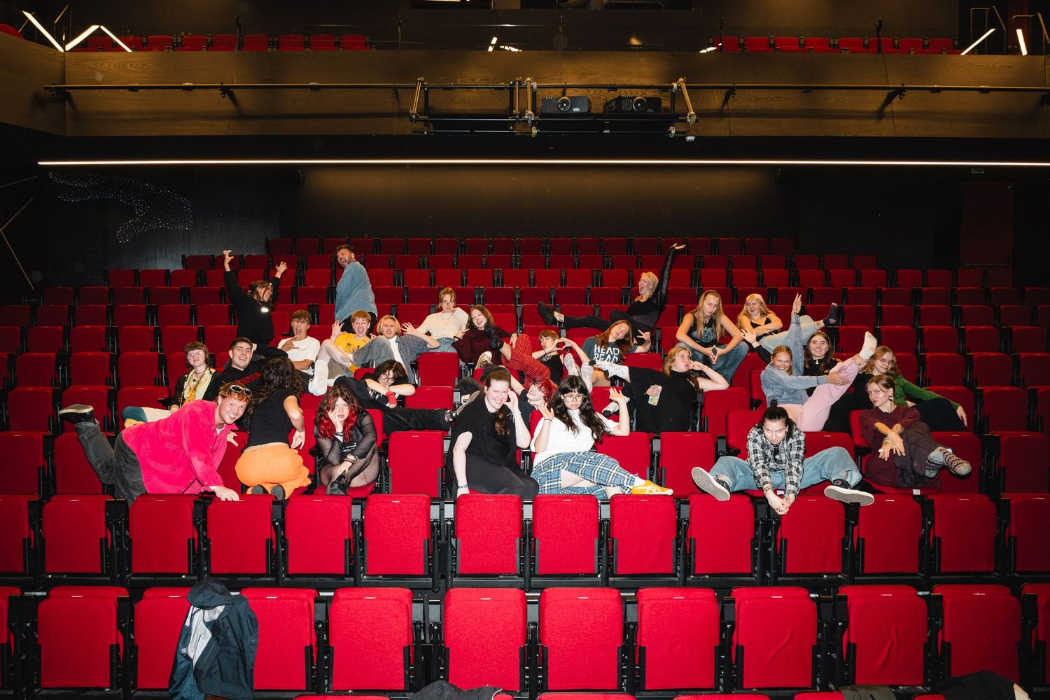 Audience members seated in red theater chairs raise their arms in excitement.