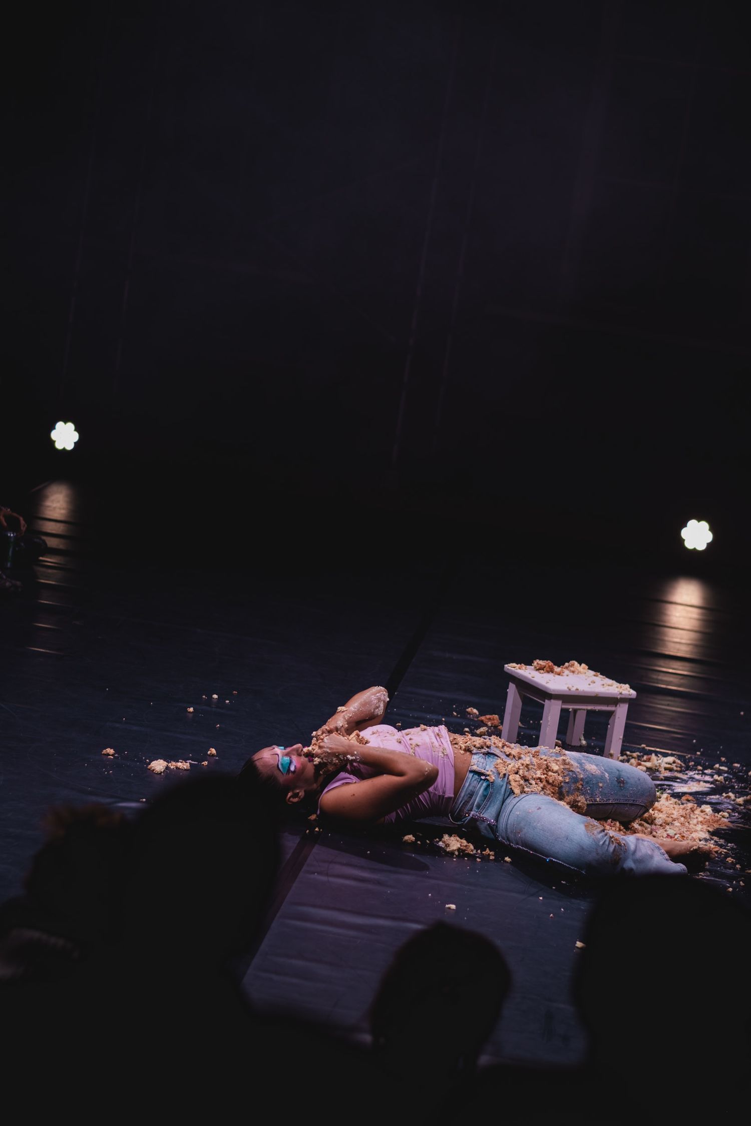 Figure lying on a dark stage surrounded by scattered materials in dramatic lighting.