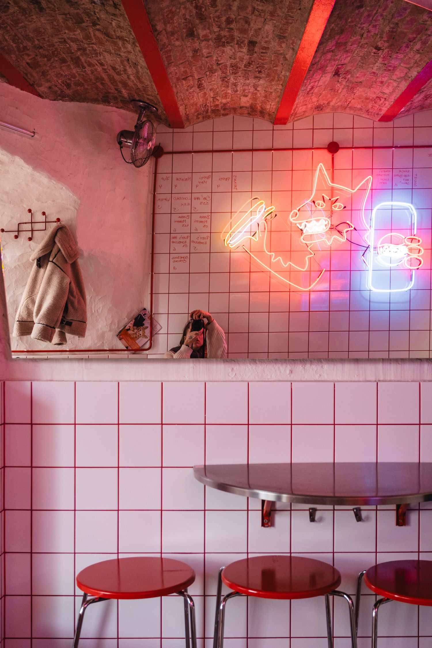 Retro diner interior with pink tiled walls and neon cat sign casting a red glow.