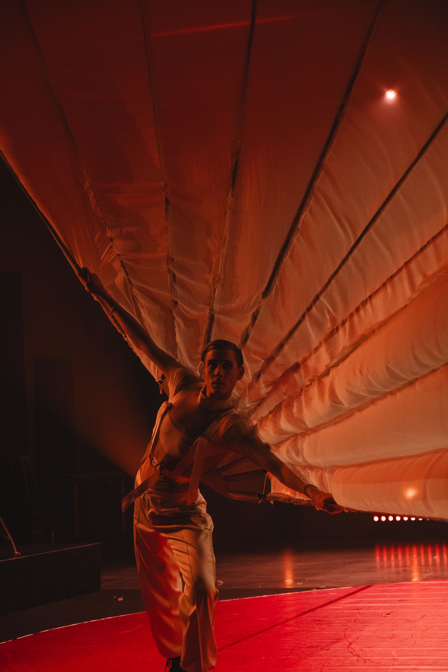 Performer in flowing white costume creating dramatic shapes under red stage lighting.