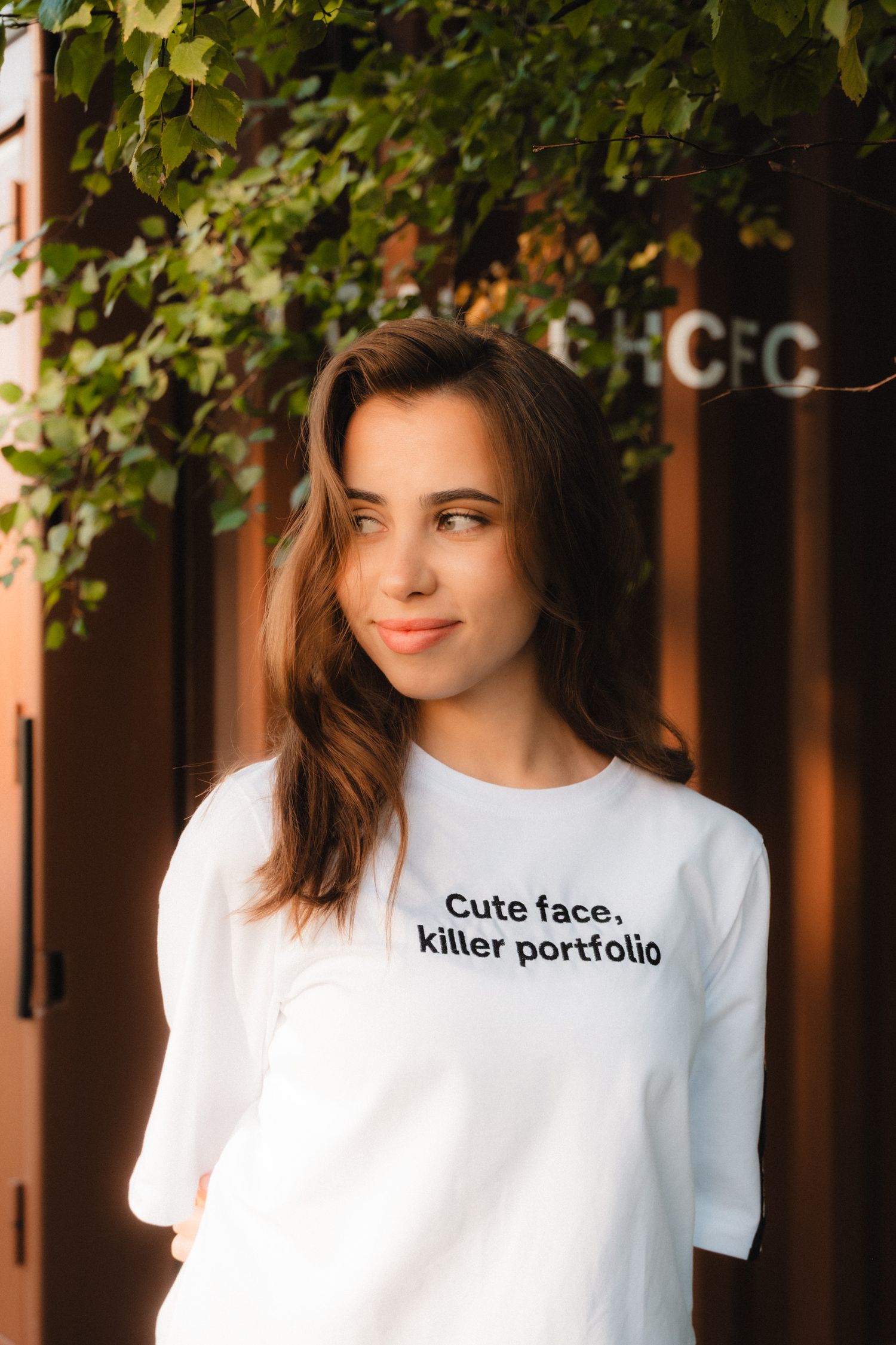 Person wearing a white t-shirt with text smiles while posing against a background with green foliage.