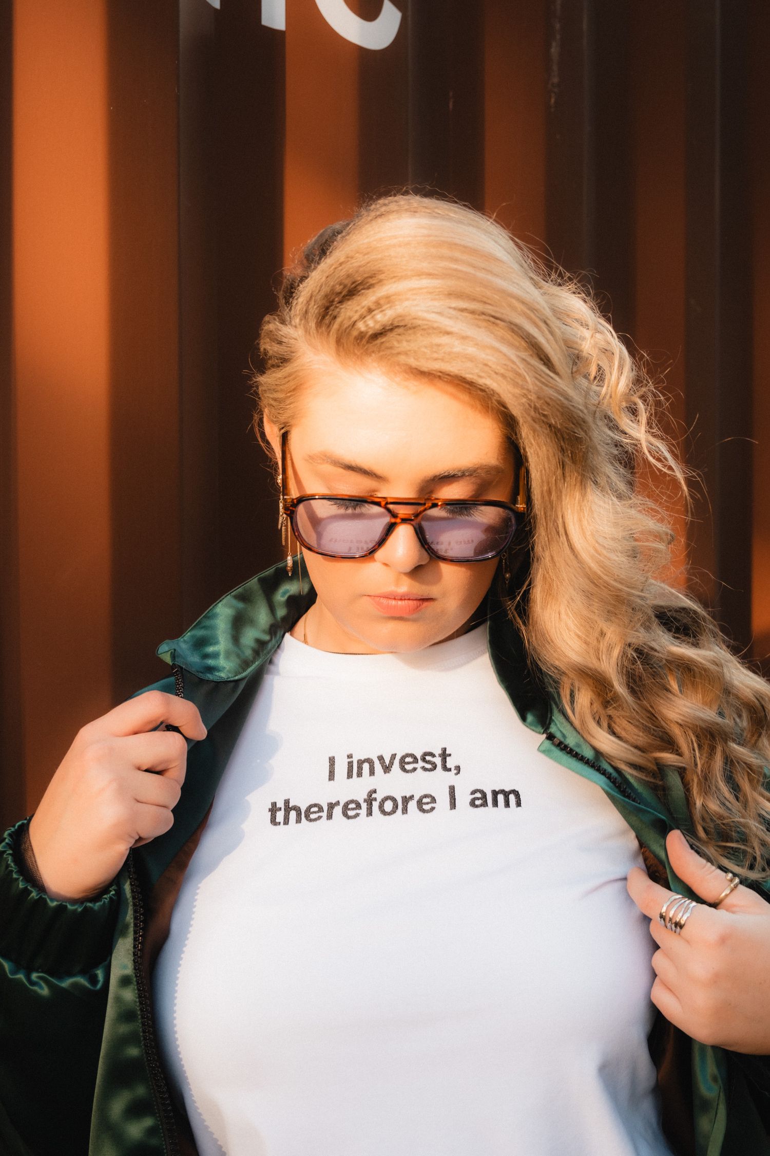 Person wearing sunglasses and a green jacket with a t-shirt that reads 'I invest, therefore I am' in an orange-lit setting.