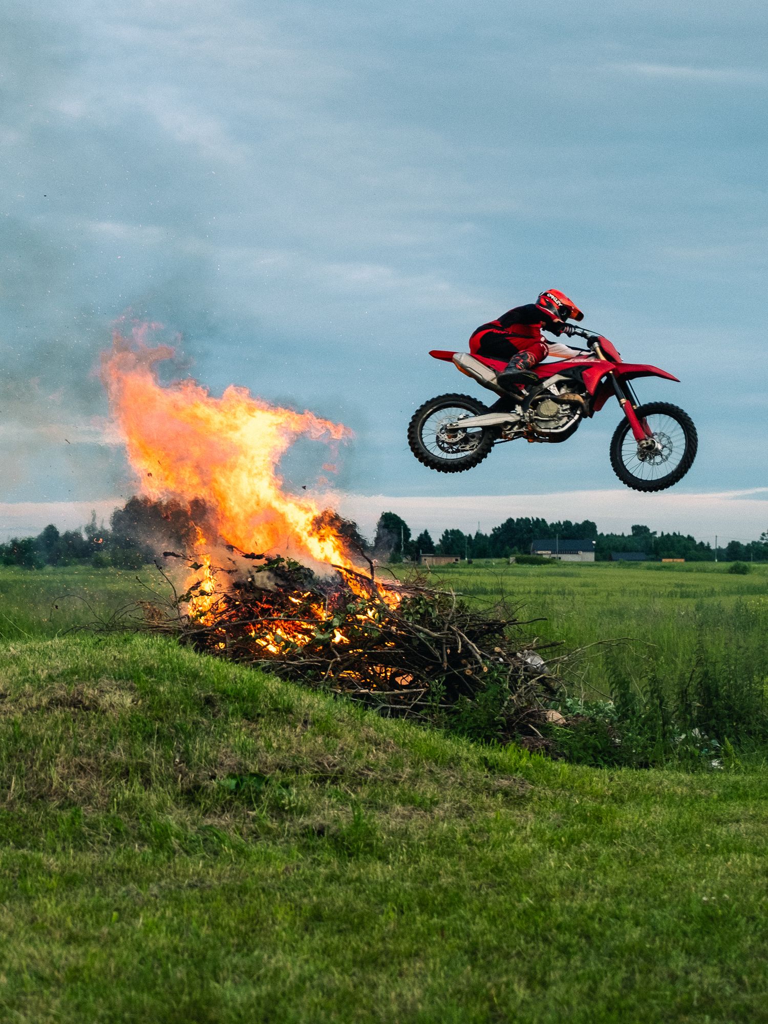 Motocross rider jumps over a large bonfire on a green field, performing a daring stunt on a red dirt bike.