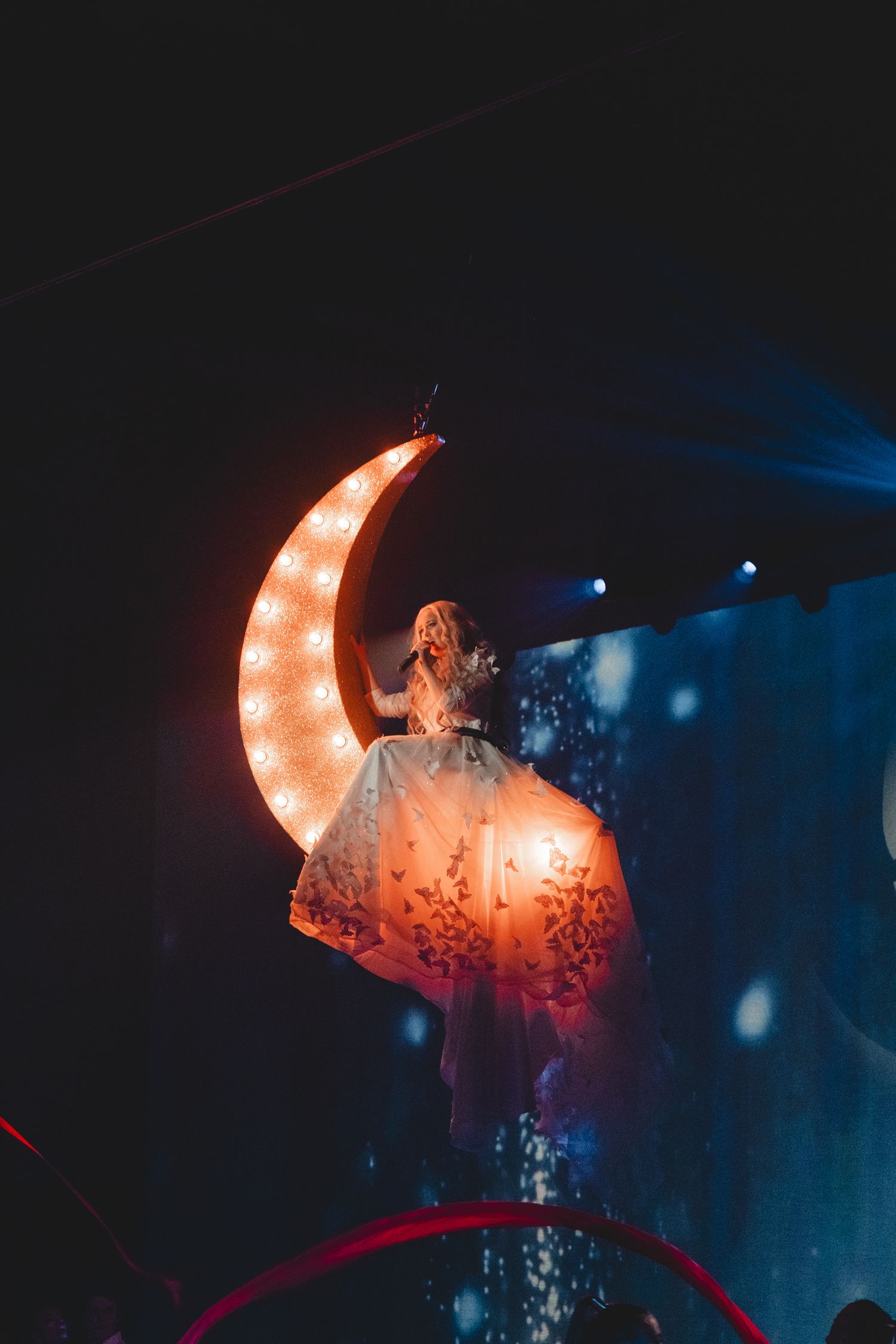 Illuminated crescent moon prop glowing orange against dark blue stage lighting.