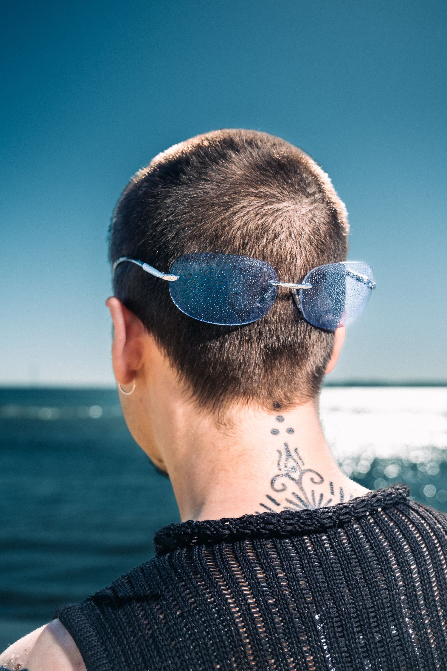 Back view of person with short hair, blue aviator sunglasses, and neck tattoo overlooking ocean on sunny day.