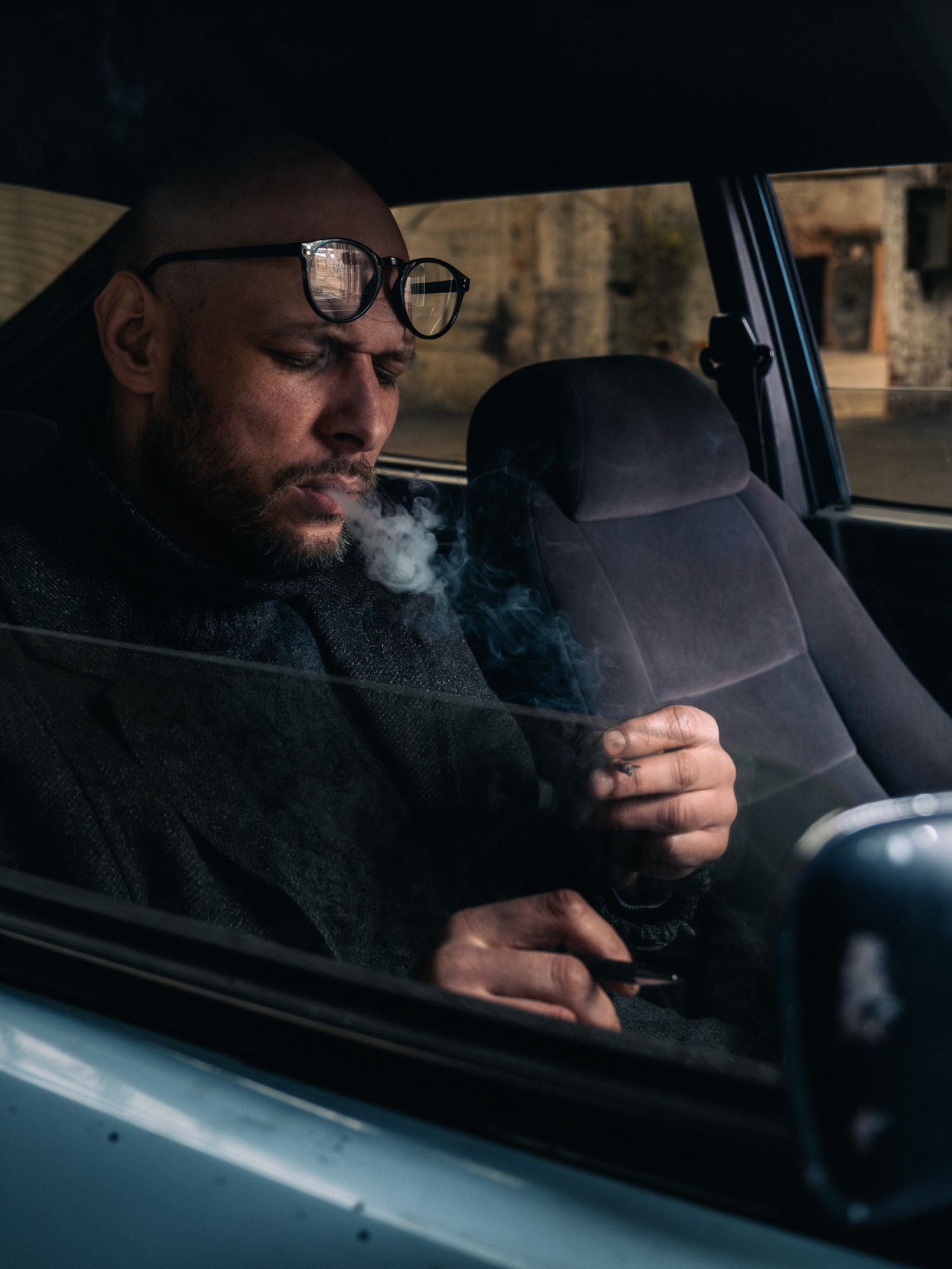 Bald man with glasses and beard smoking inside a vintage car, exhaling smoke in moody natural light.