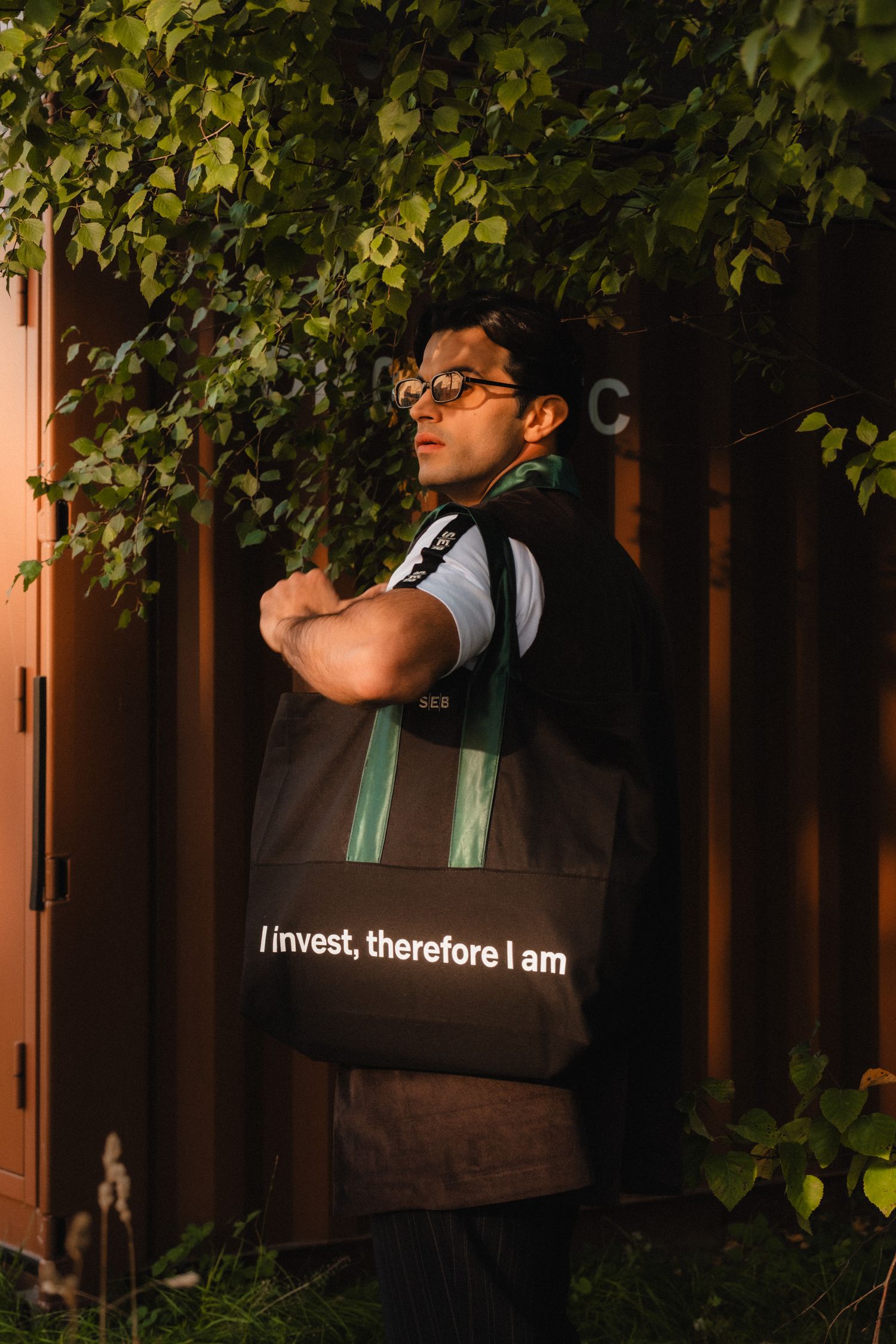 Person wearing glasses and a backpack stands against a wooden fence with green foliage in the background.
