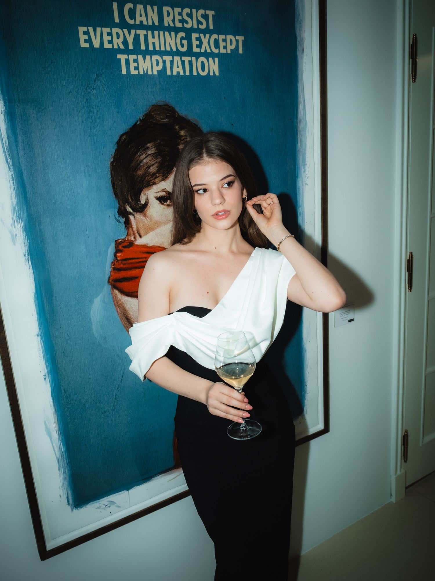 Figure in black and white outfit holds martini glass against blue wall with text reading 'I Can Resist Everything Except Temptation'.