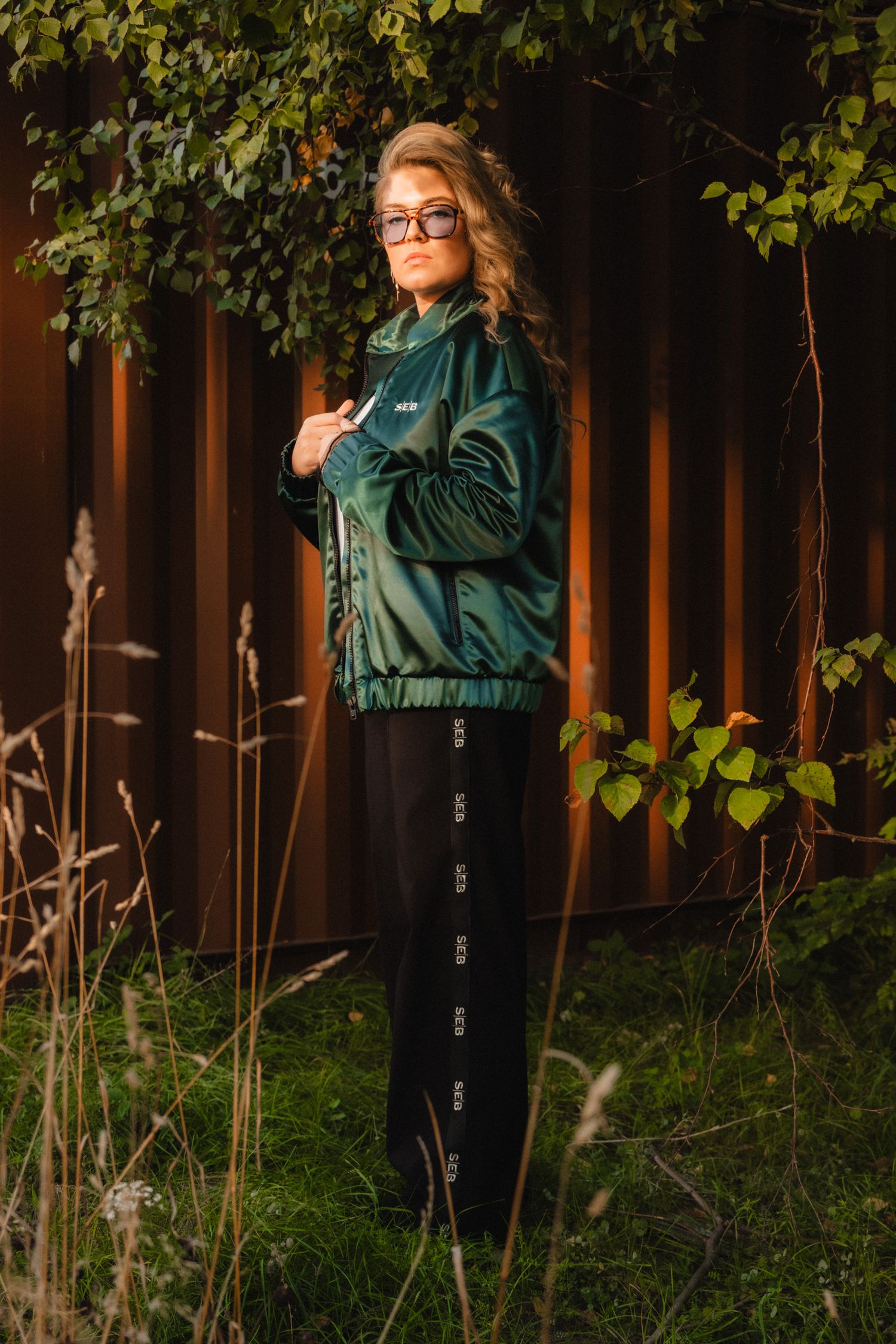 Person in green jacket and glasses poses in front of wooden fence with climbing vines in backyard garden setting.