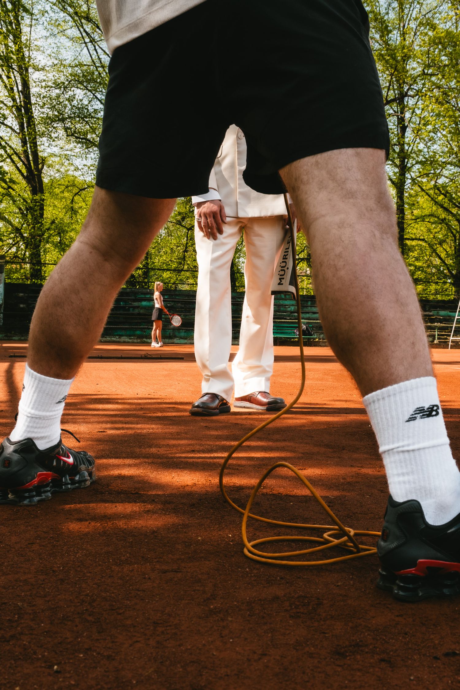 Close-up of tennis players' legs on a clay court, with New Balance socks and a racket visible in background.