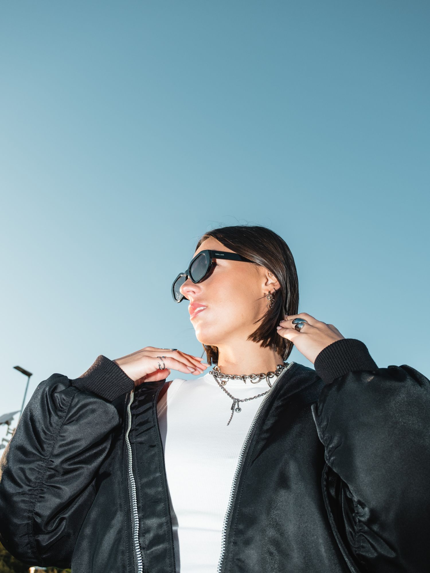 A person in sunglasses and black bomber jacket poses against a blue sky background.