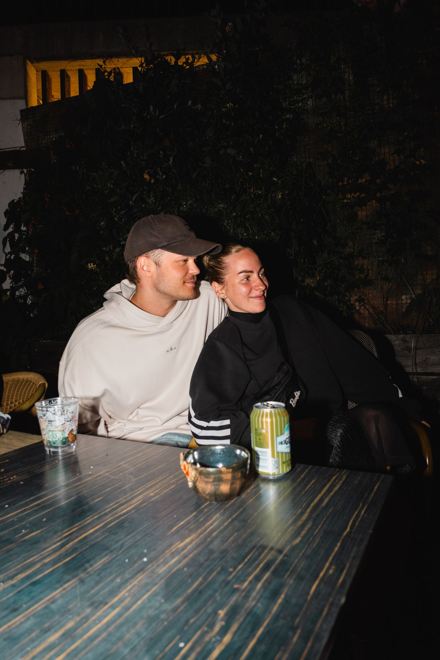 People sitting at a wooden table with drinks in a dimly lit outdoor setting.