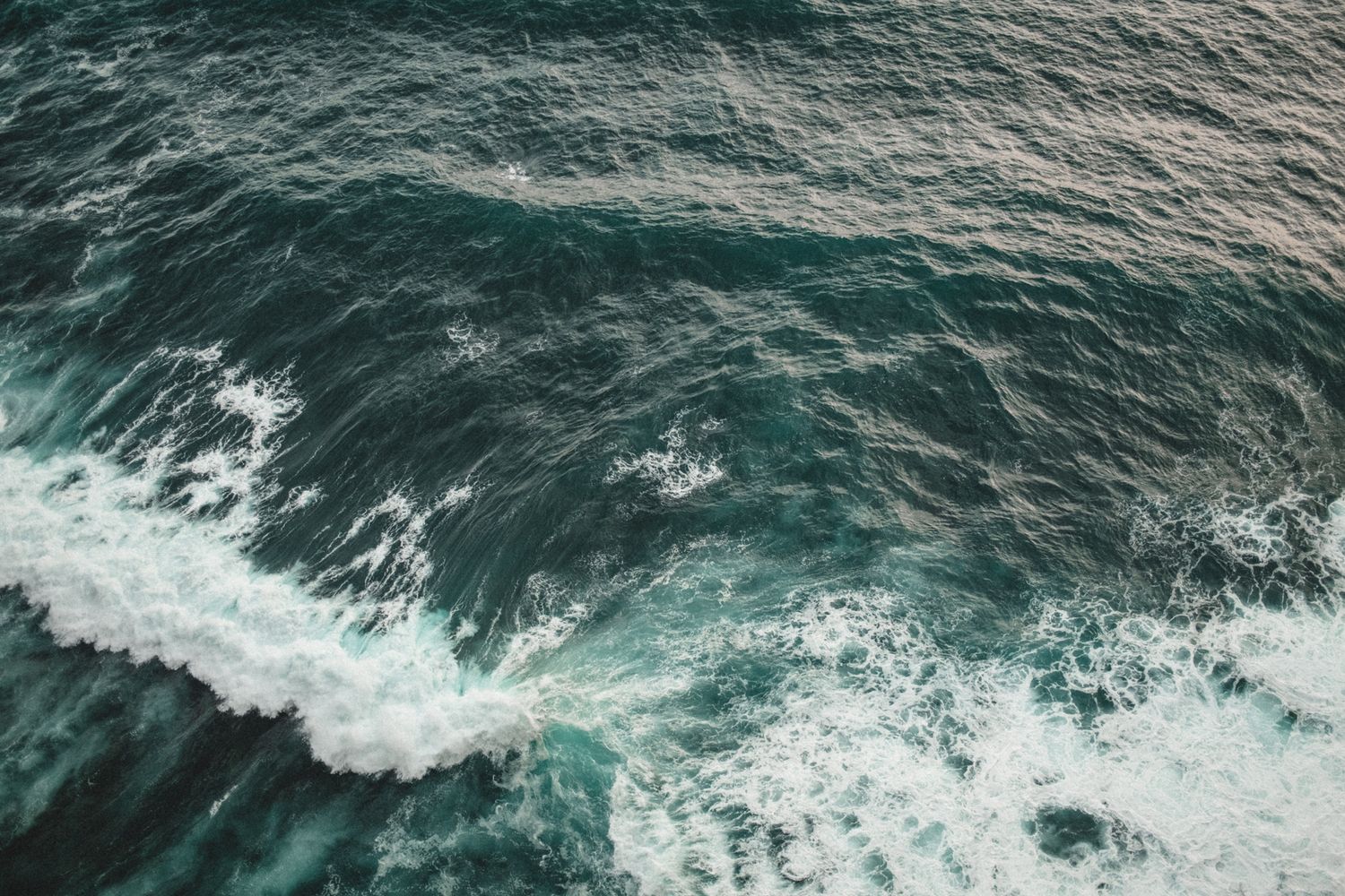 Aerial view of turquoise ocean waves crashing against rocky cliffs on a cloudy day.
