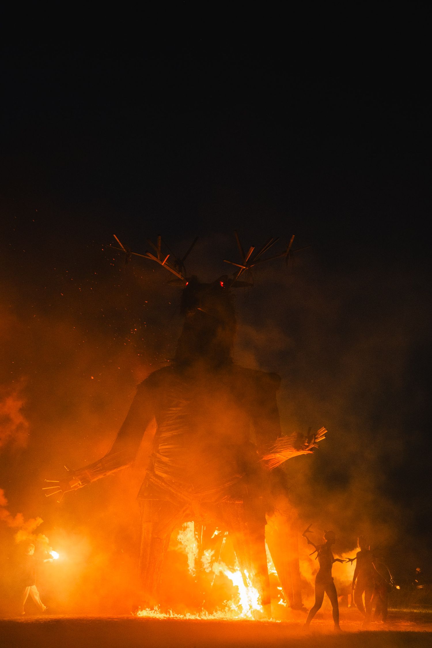 Fiery performance art with silhouettes against orange flames and smoke in darkness.