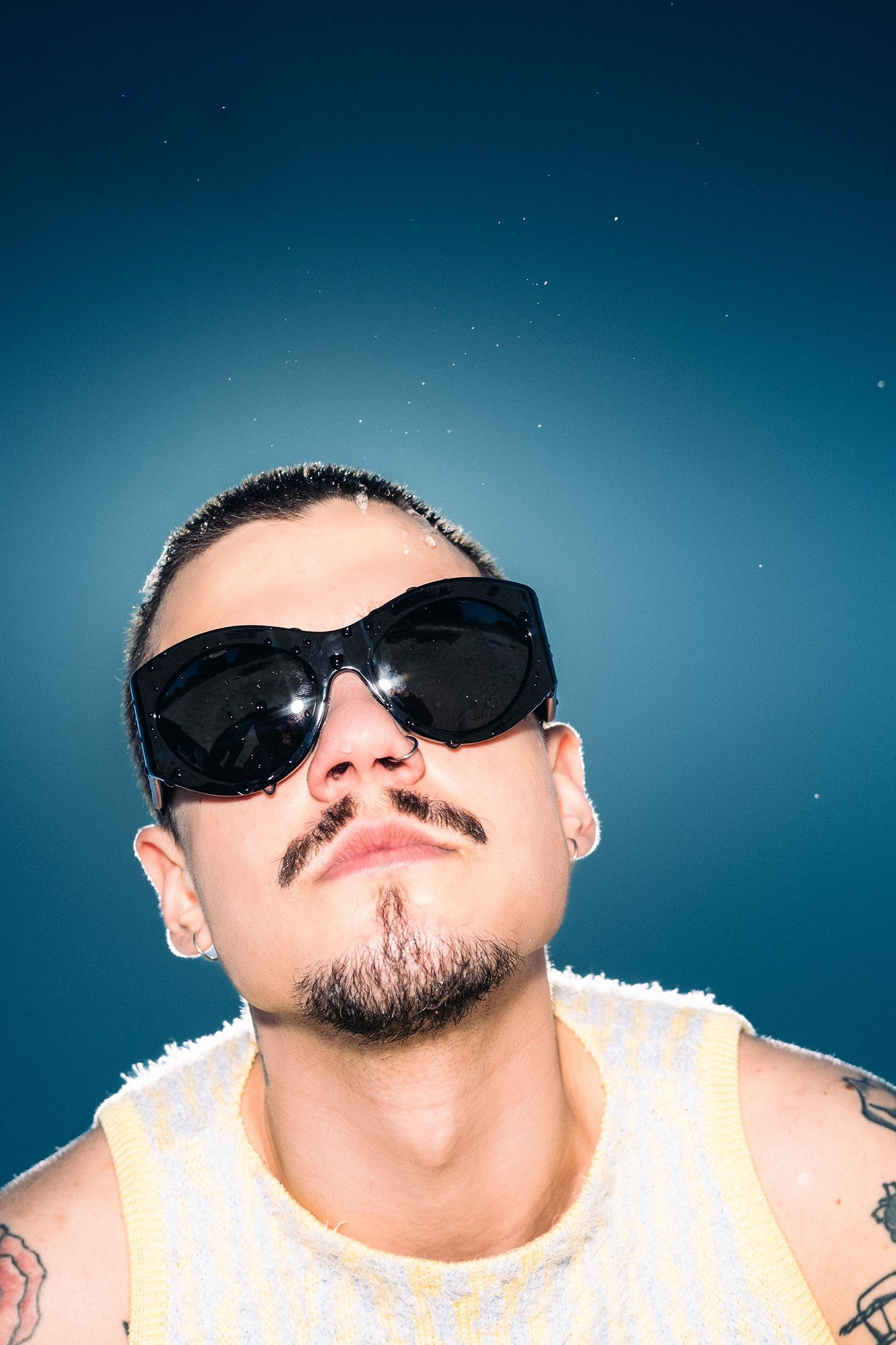 Tattooed man with goatee wearing large black sunglasses and white tank top poses against blue background.