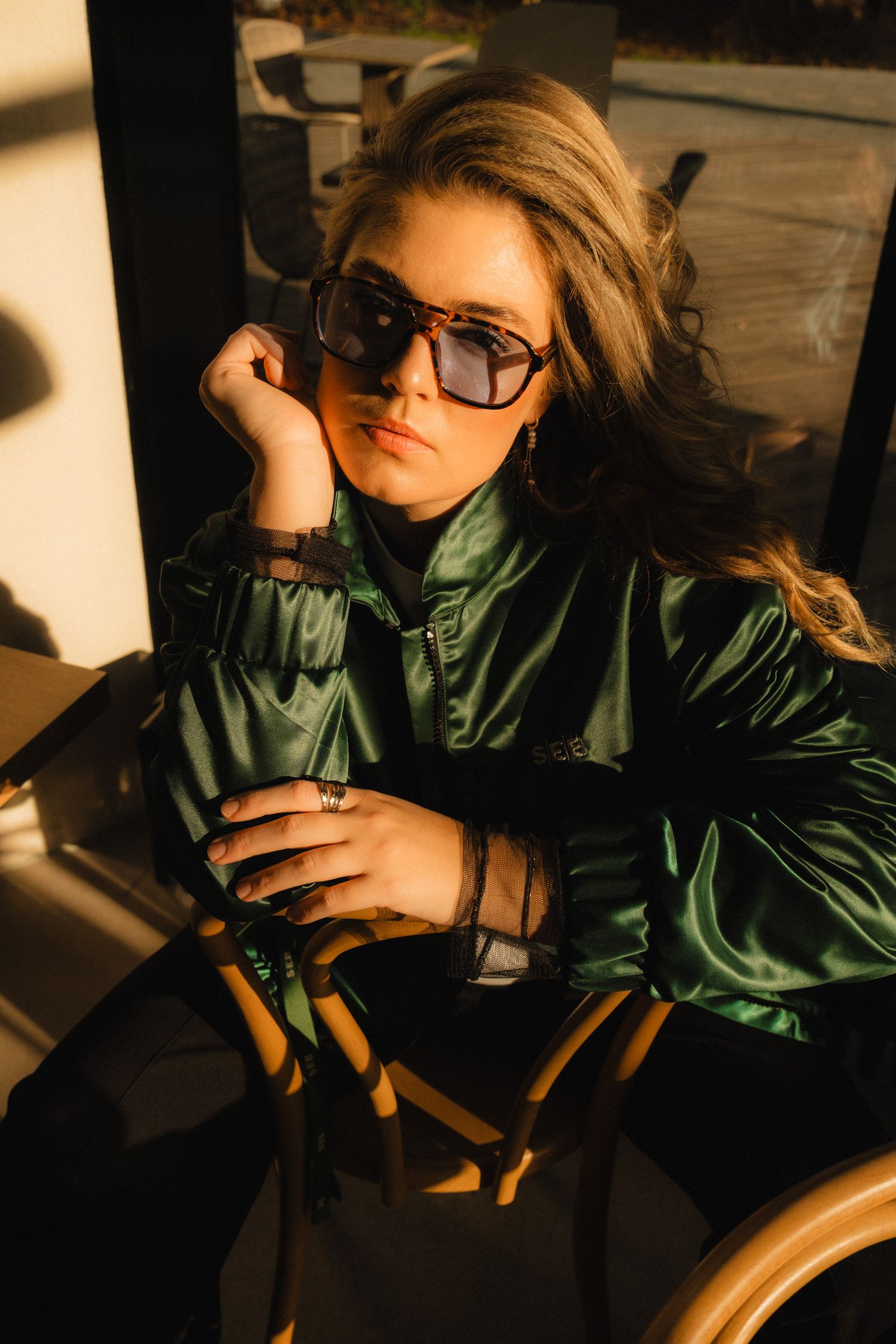 Stylish figure in sunglasses and shiny green puffer jacket poses in warm lighting against wooden furniture.