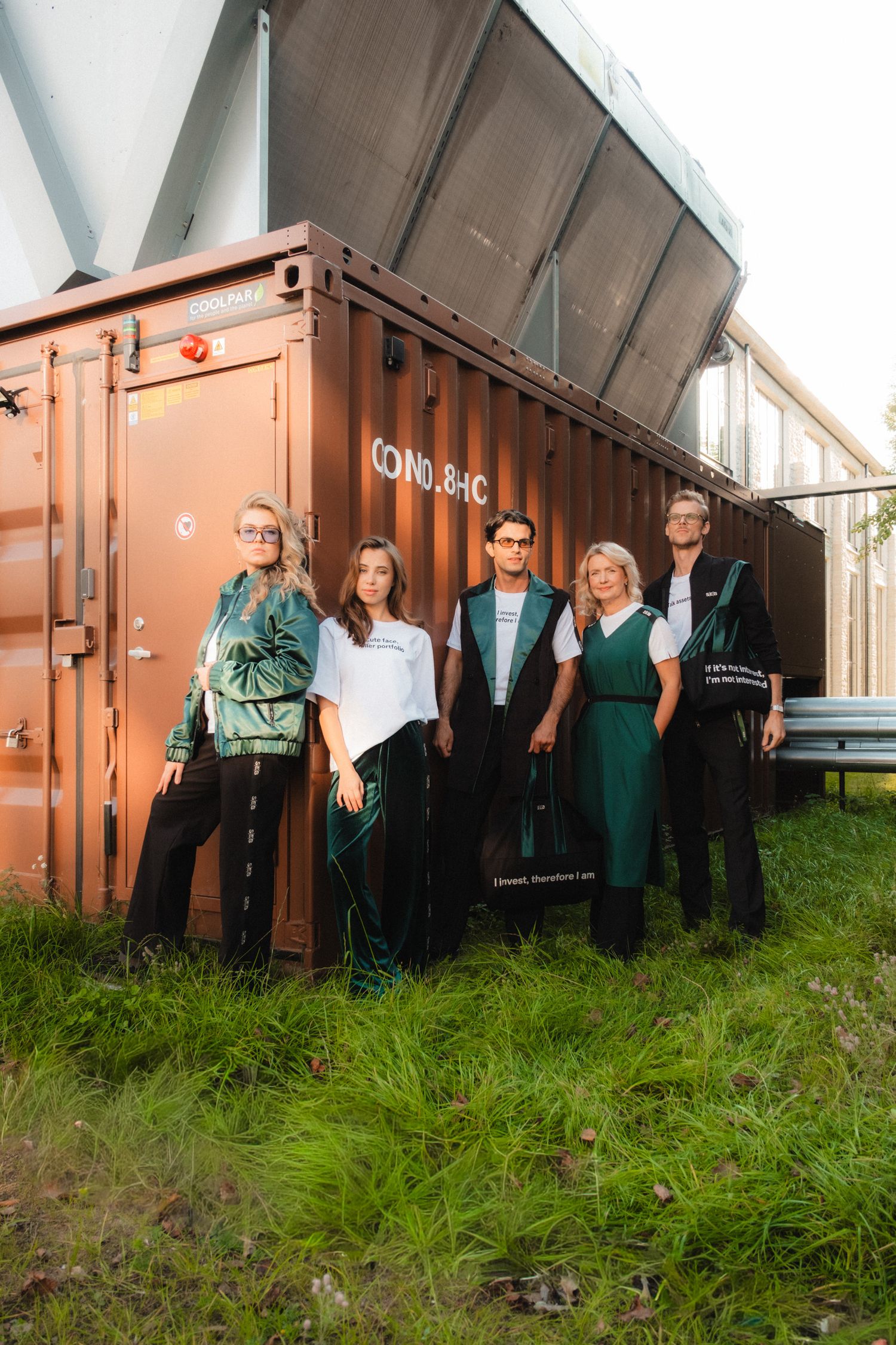 Group stands in front of orange shipping container on grassy area near industrial building in outdoor setting.