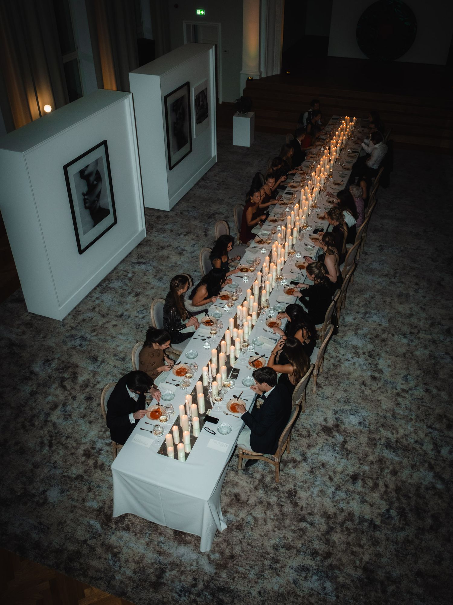A long white dining table with lit candles creates an intimate atmosphere in a dimly lit gallery space.