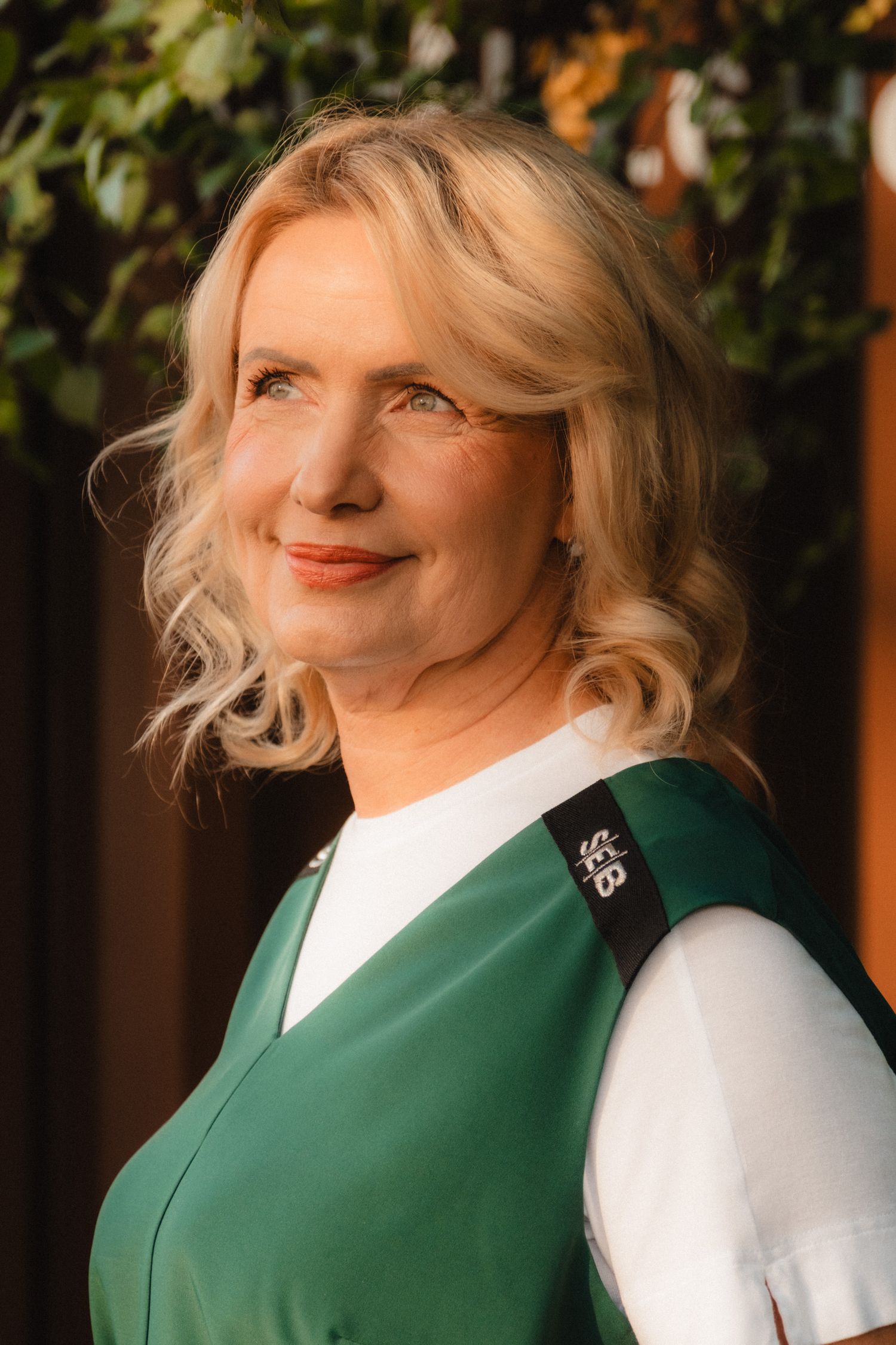 A person wearing a white shirt and green vest smiles warmly in front of foliage during golden hour lighting.